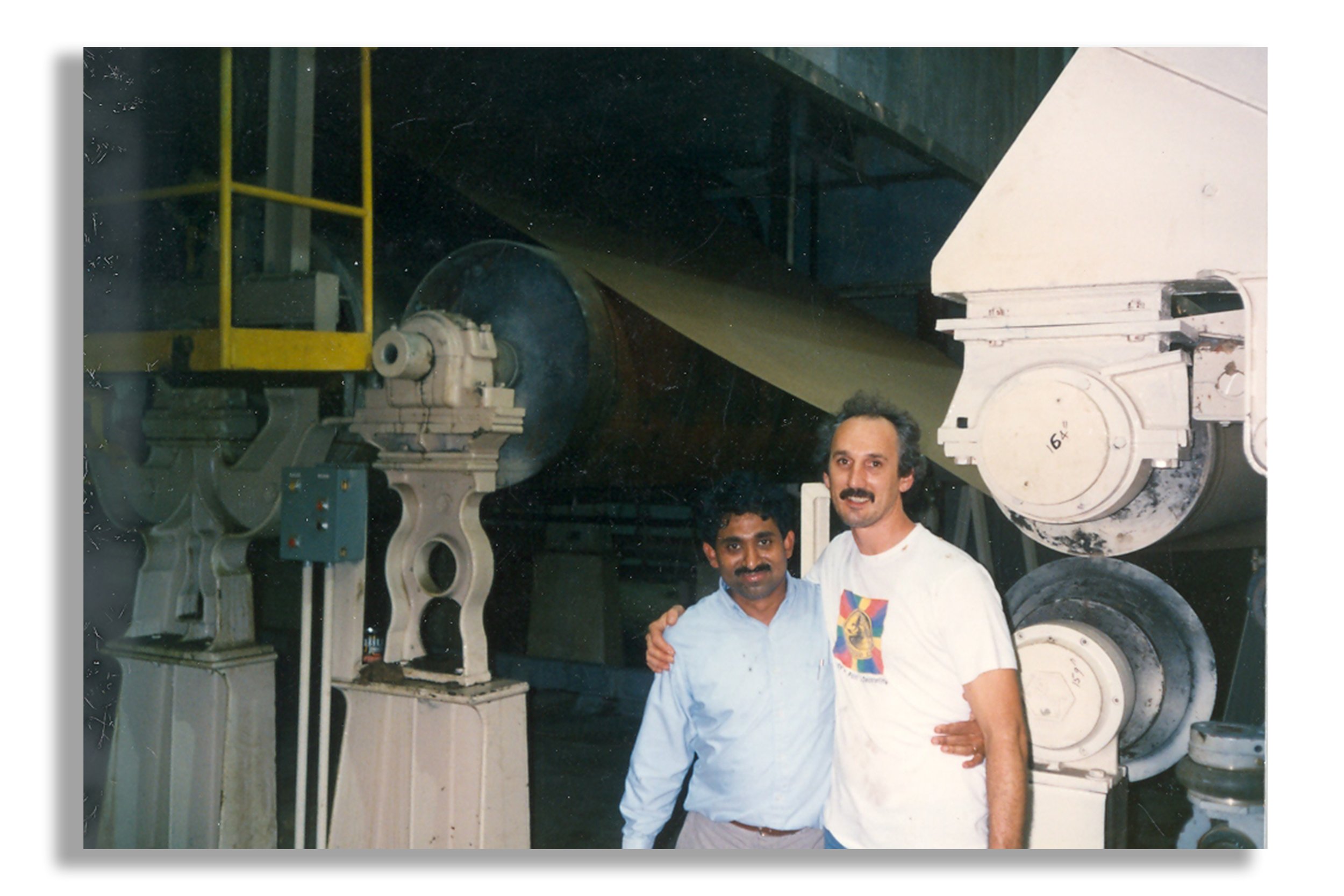 Two men standing together in front of large industrial machinery in a factory or industrial setting, with one man's arm around the other's shoulder.
