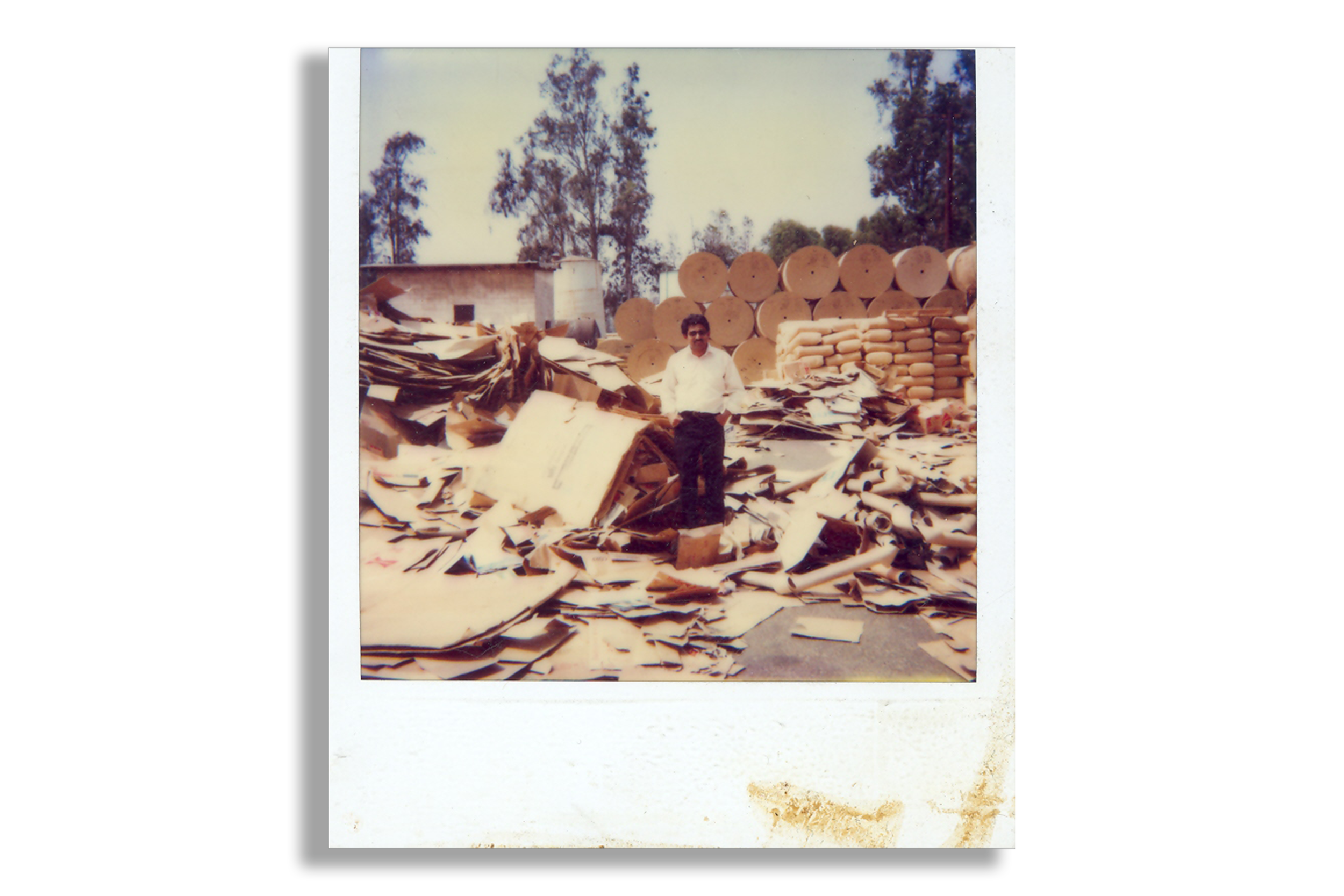 A man standing on a pile of cardboard and paper scraps in an industrial yard with stacks of large cardboard rolls and bags behind him, and trees in the background.
