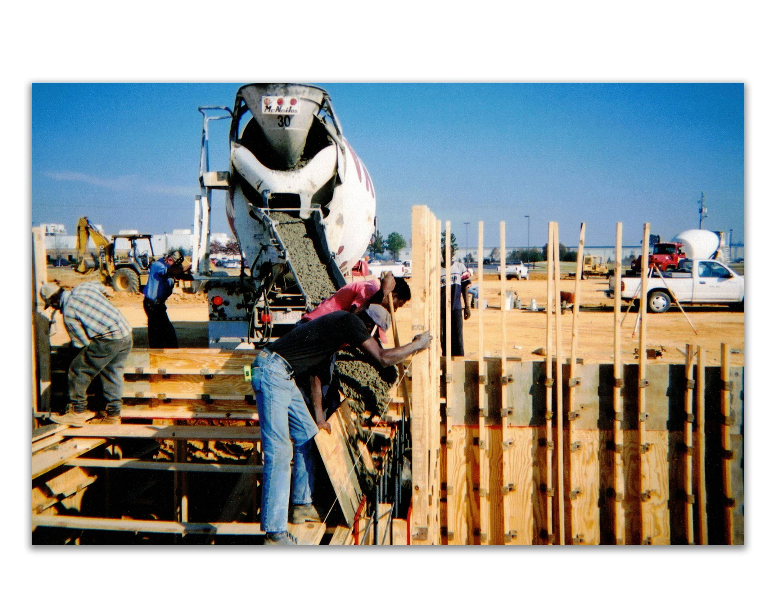 Construction workers pouring concrete at a building site with wooden framework, a cement mixer truck, and construction equipment in the background.