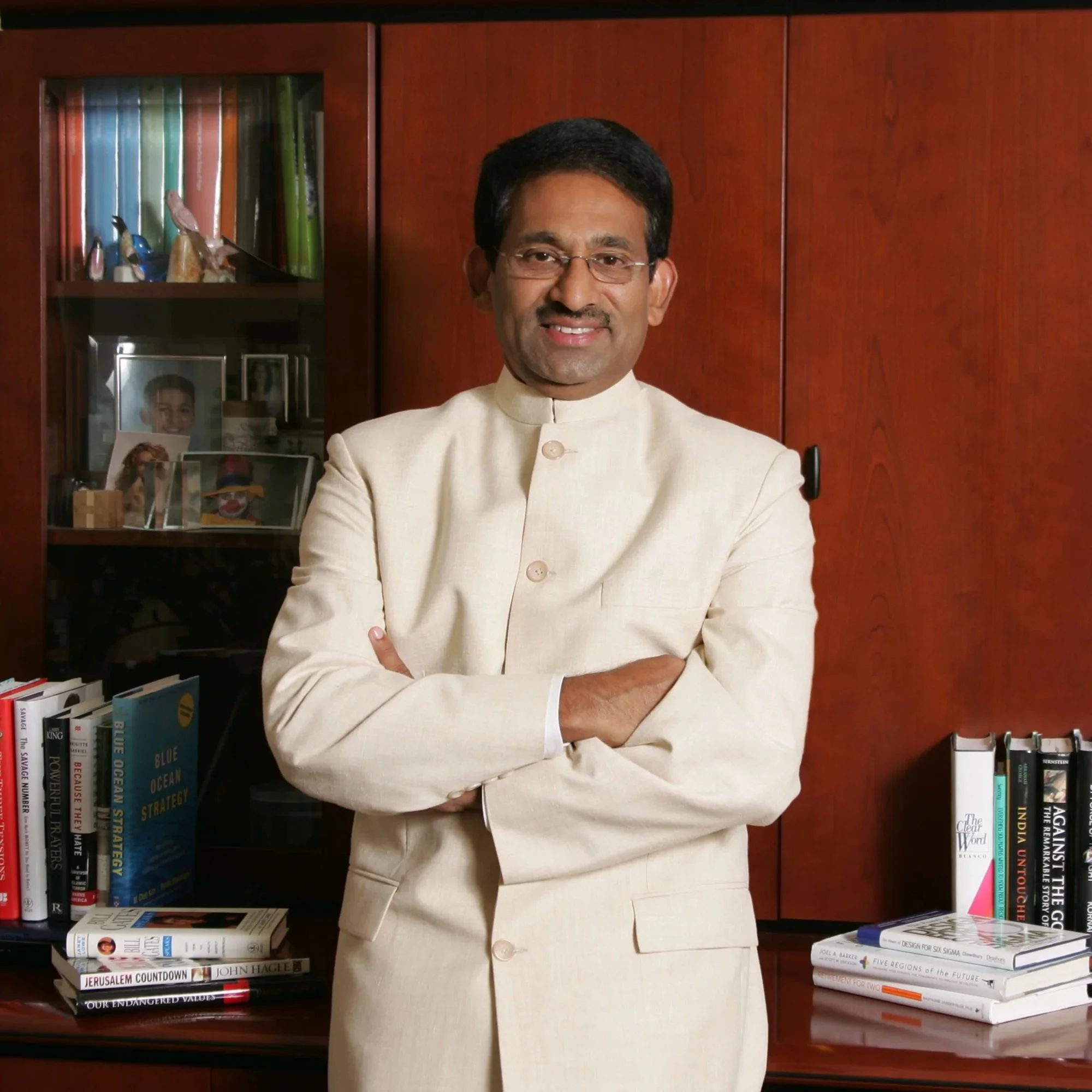A man in a cream-colored traditional Indian suit standing with arms crossed in an office. Behind him is a bookshelf with books and family photos, and a wooden wall.