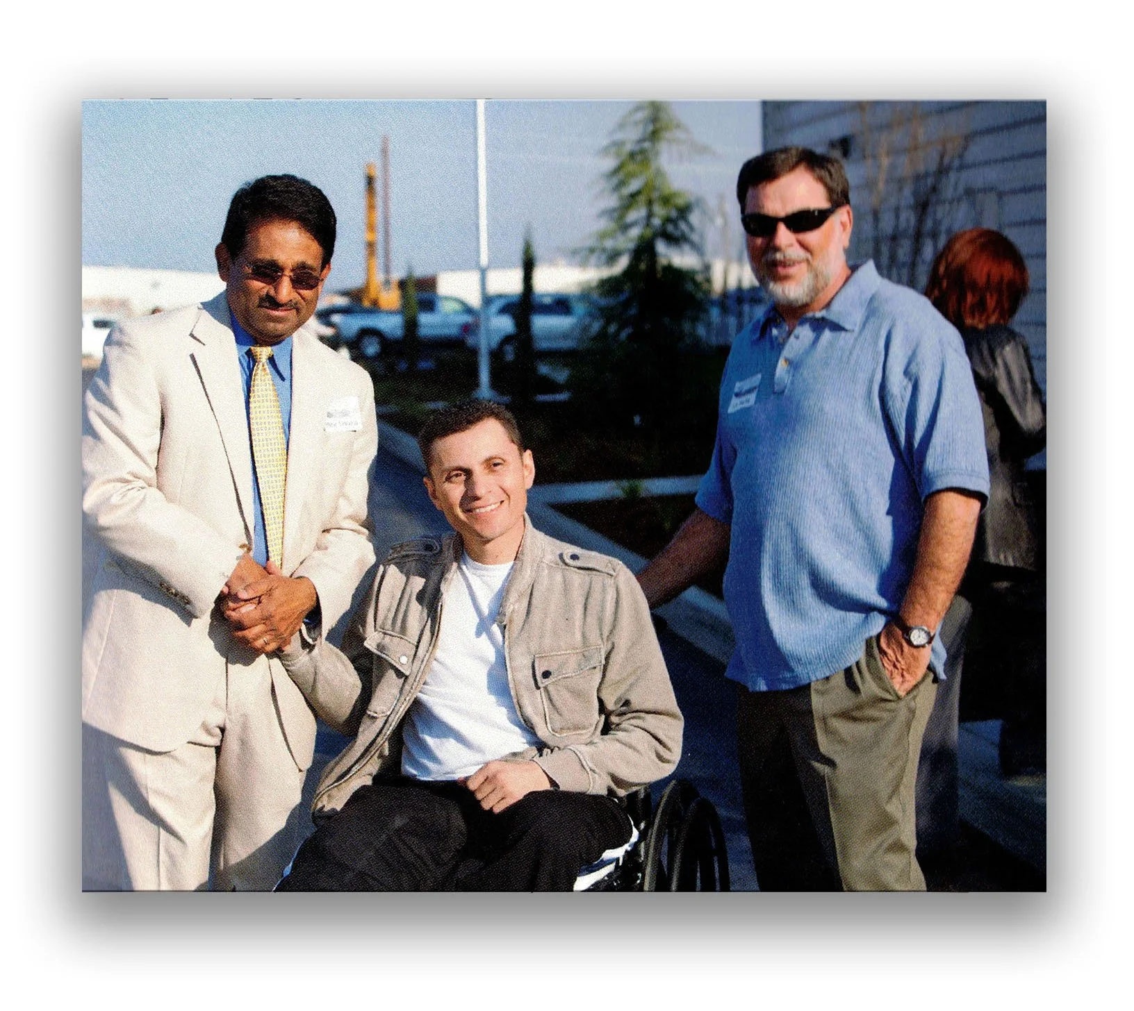 Three men, one in a wheelchair, standing outdoors in a parking lot during daytime, smiling at the camera. All are dressed casually or in business attire, with two wearing sunglasses.