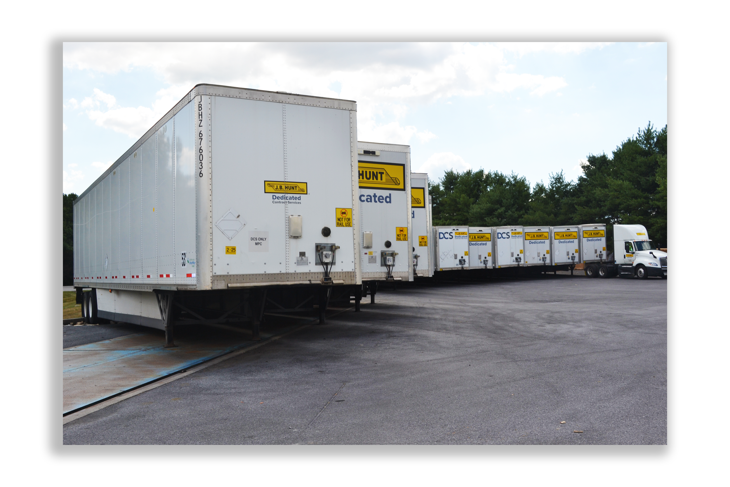 Multiple white semi-trailer trucks parked in a lot, with trees and a partly cloudy sky in the background.