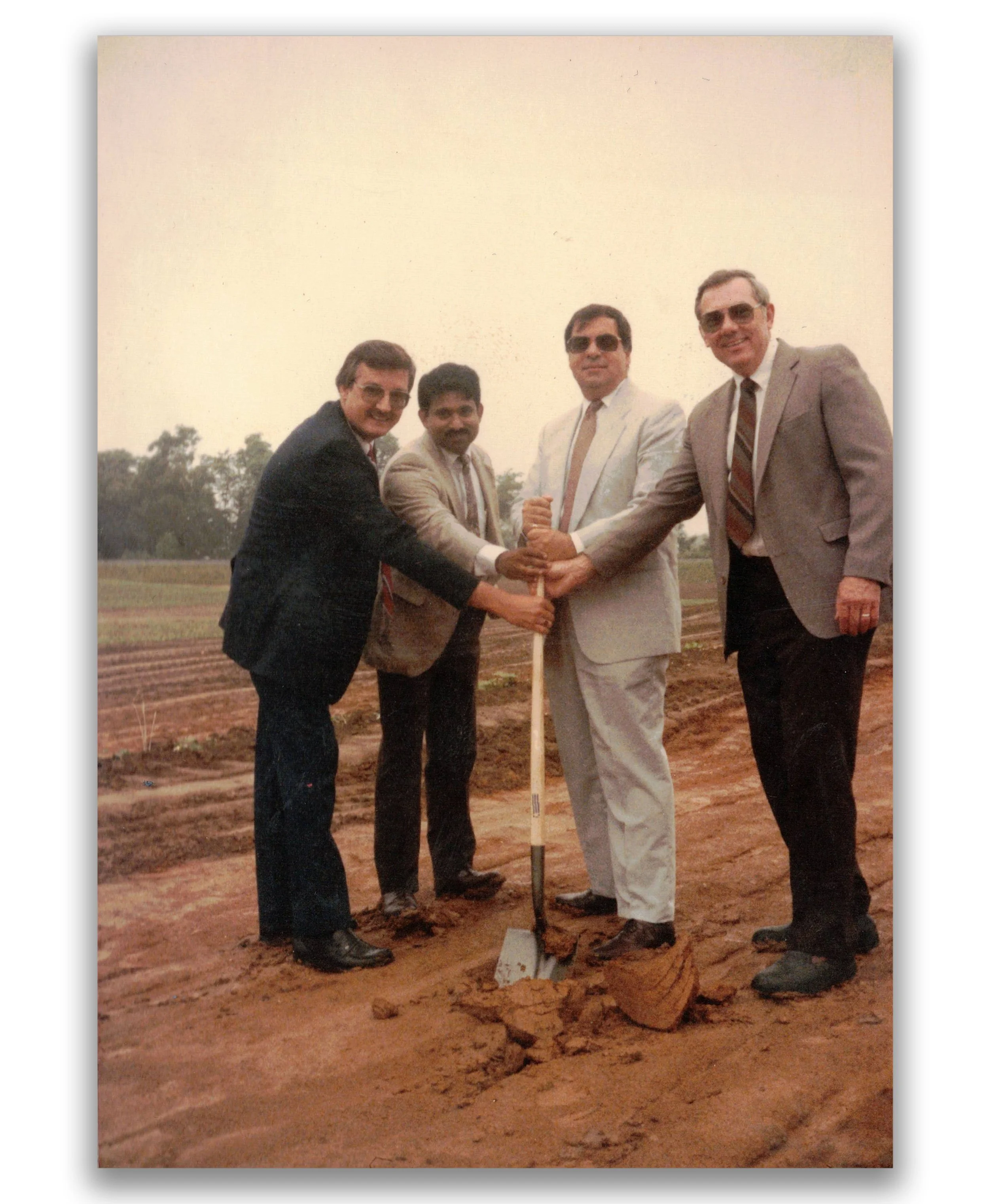 Four men in suits and sunglasses, associated with a groundbreaking or planting event, are standing on a dirt field with a shovel, holding a small mound of dirt together. One man is holding a shovel upright, and they are all smiling.
