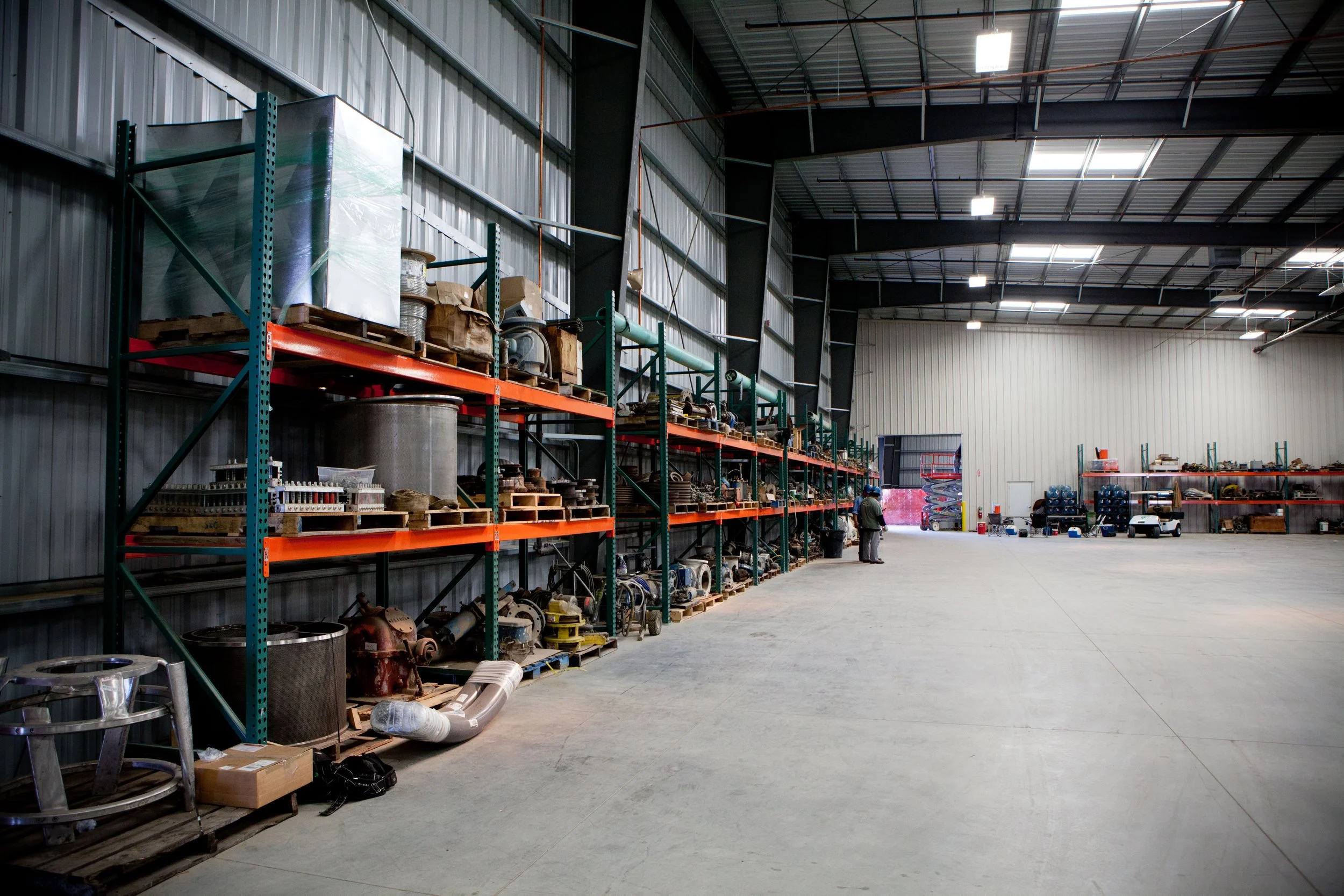 Interior of a large industrial warehouse with metal shelves holding machinery parts and equipment along the left wall, and a wide open space with a couple of people near the background door.
