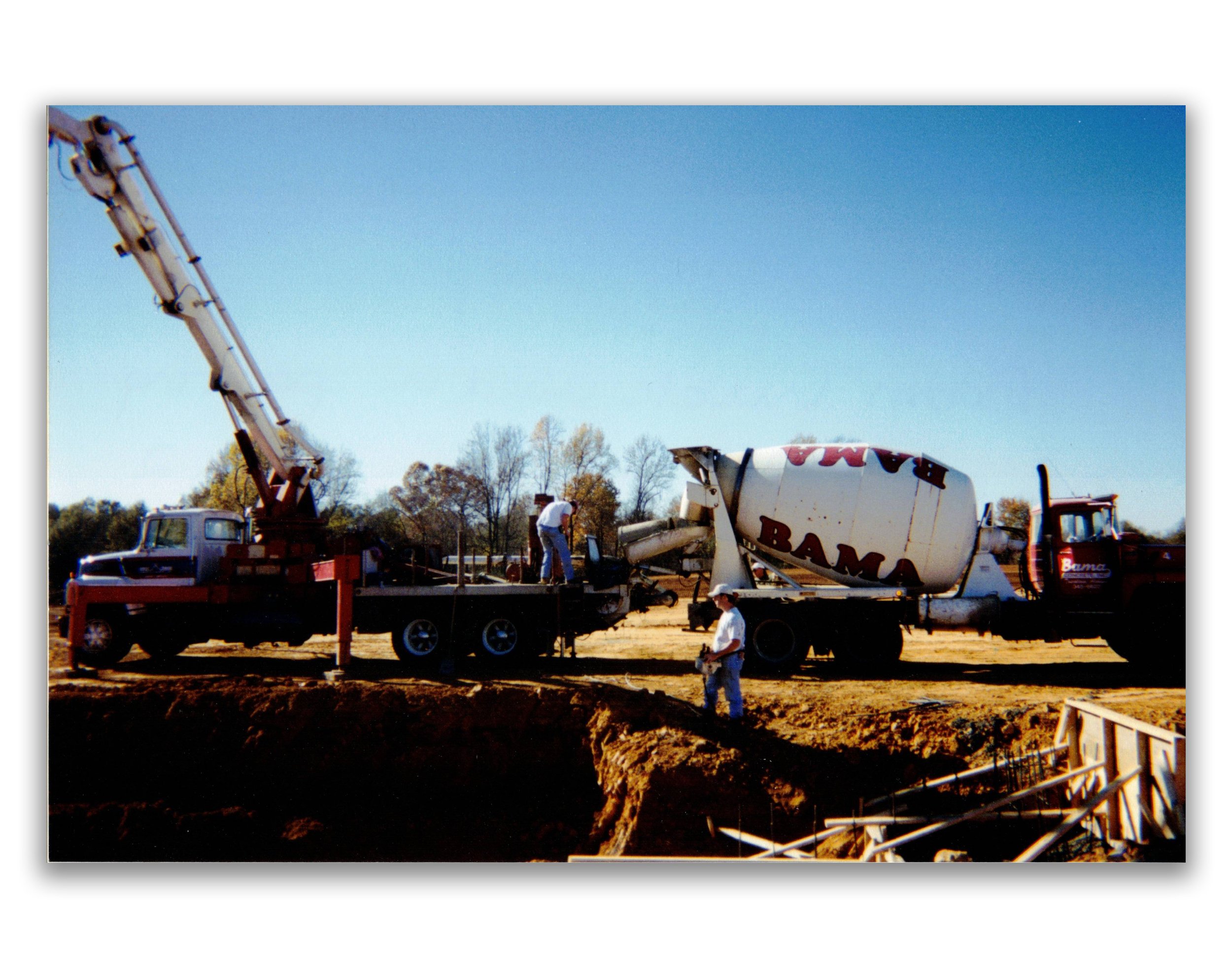 Construction site with a cement mixer truck and workers, one operating the truck's crane and another on the ground, under a clear blue sky.