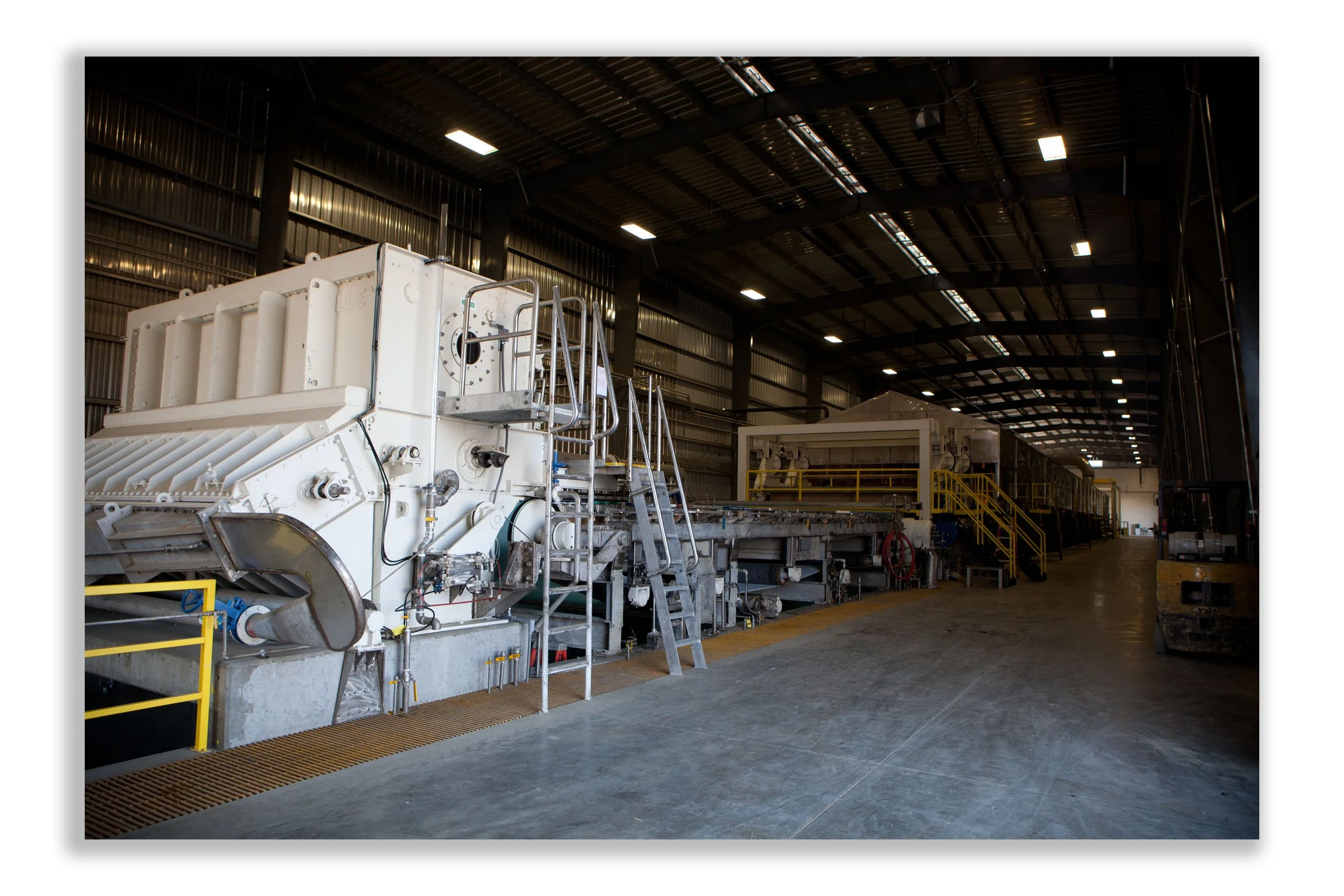 Interior of an industrial warehouse with large machinery, metal stairs, yellow safety railings, and overhead lighting.