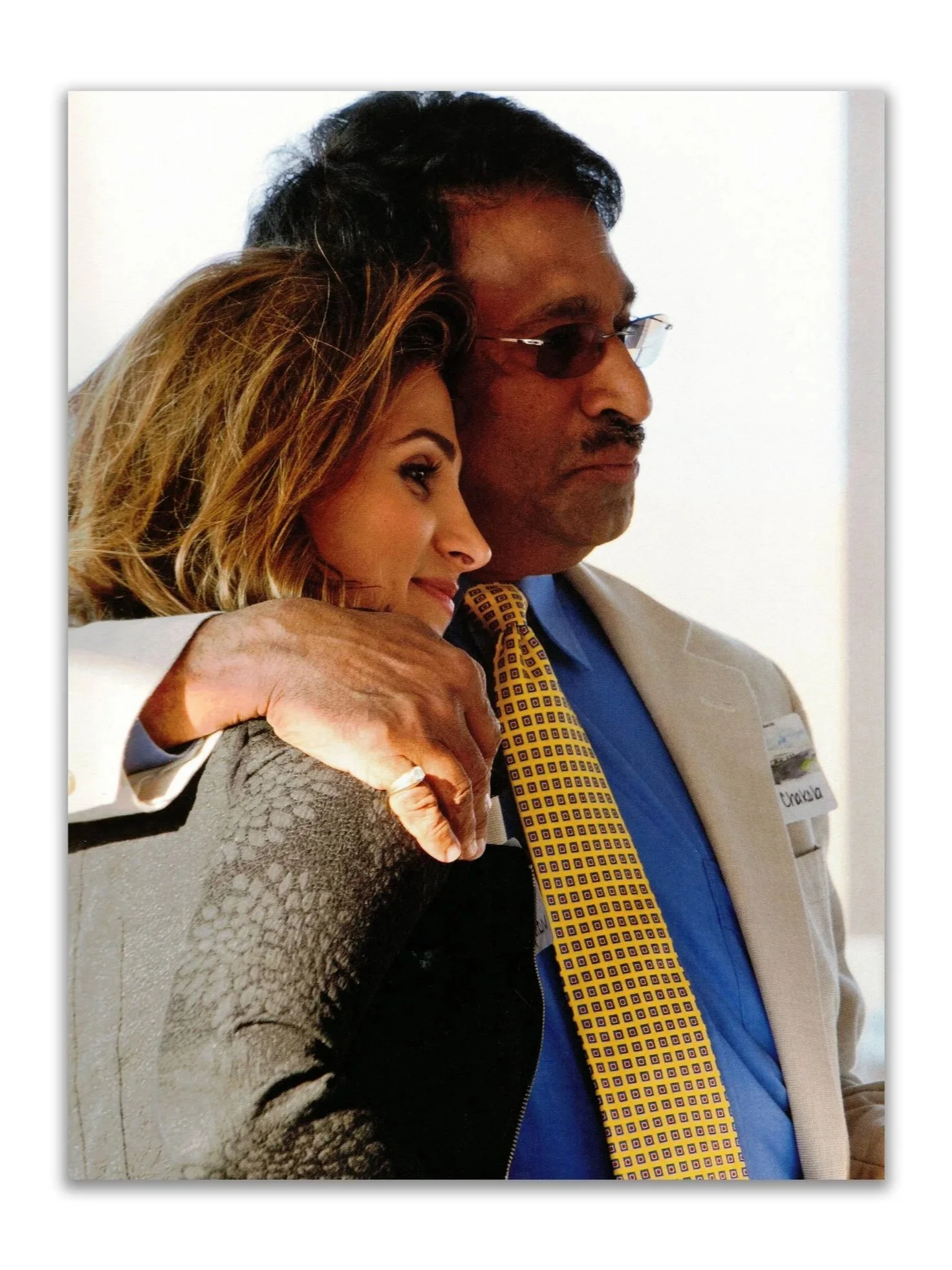 A woman and a man in a friendly pose, with the woman leaning on the man’s shoulder, both looking to the side. The woman has light brown hair, and the man has dark hair, glasses, and is wearing a tan jacket with a blue shirt and a yellow patterned tie.