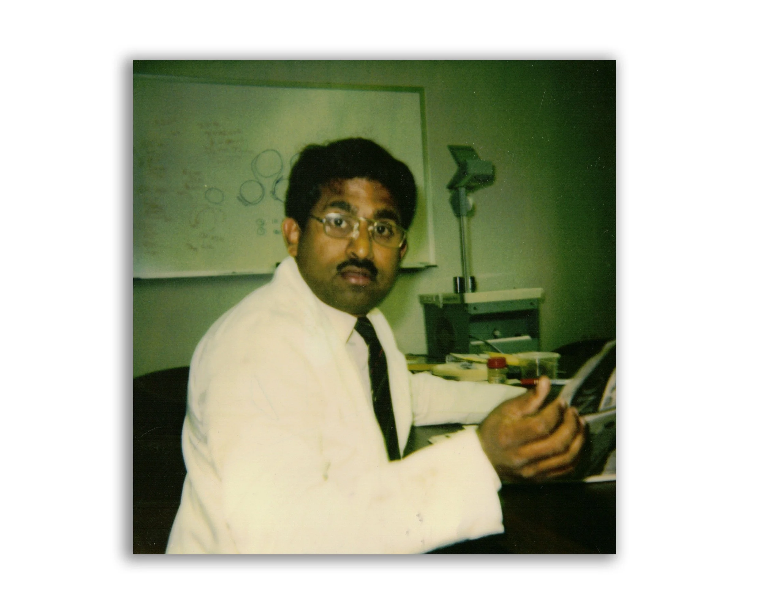 A man with glasses and a mustache, wearing a white lab coat, seated at a desk in an office or laboratory, looking at the camera. In the background, there is a whiteboard with diagrams and a laboratory instrument.