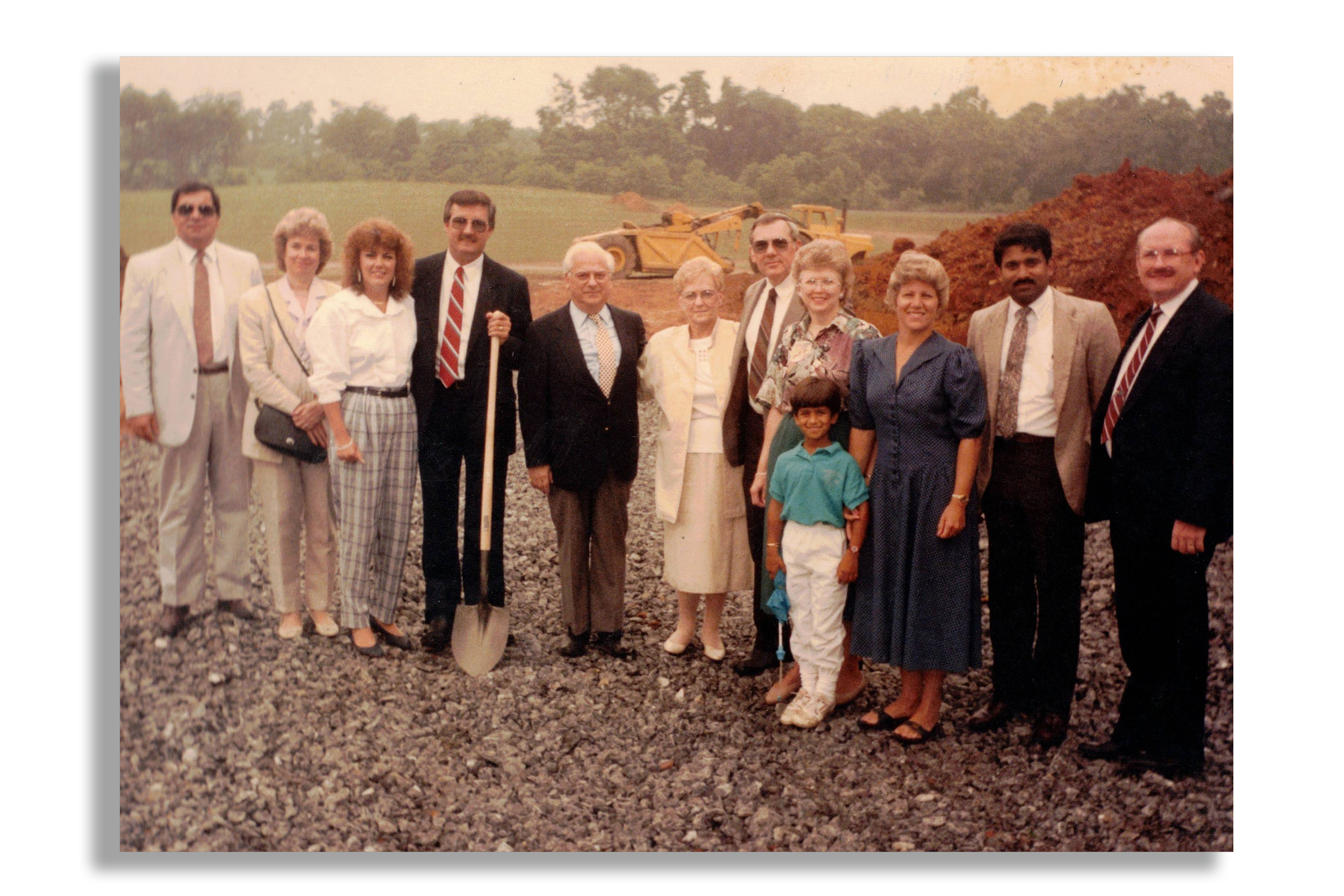 A group of about thirteen people, including men, women, and children, standing on a rocky surface outdoors in front of a green field, trees, and a yellow construction vehicle, during daytime.
