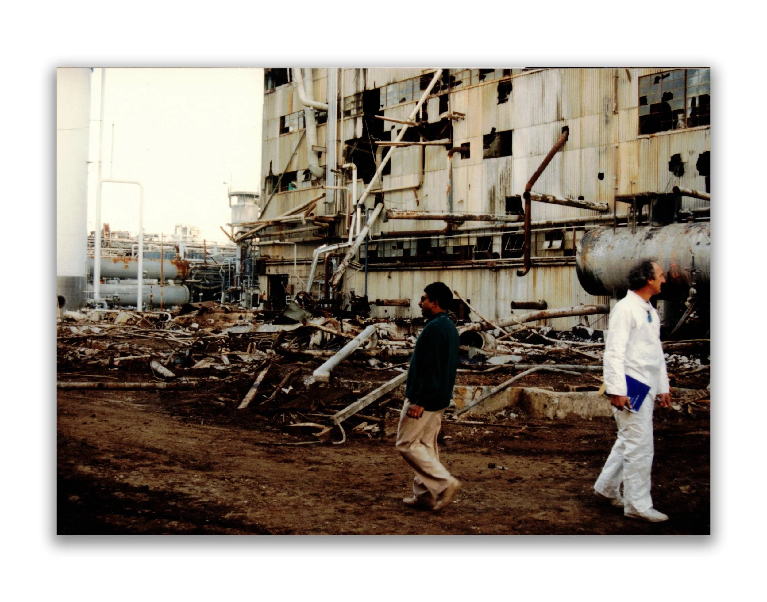 Two men walk past a damaged industrial building with debris and rusted pipes on the ground.