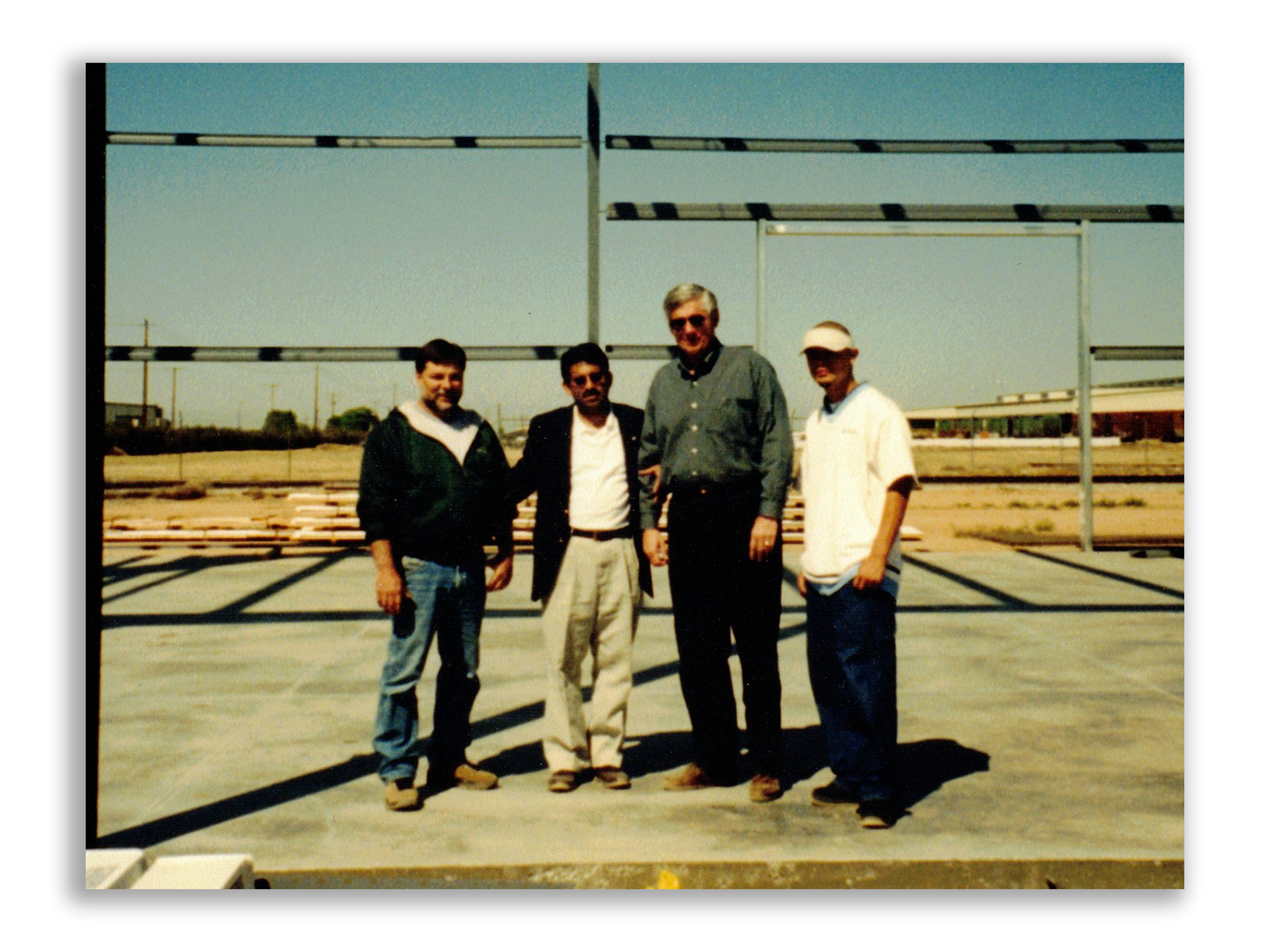Group of four men standing outdoors on a construction site with metal framing and a clear sky in the background.