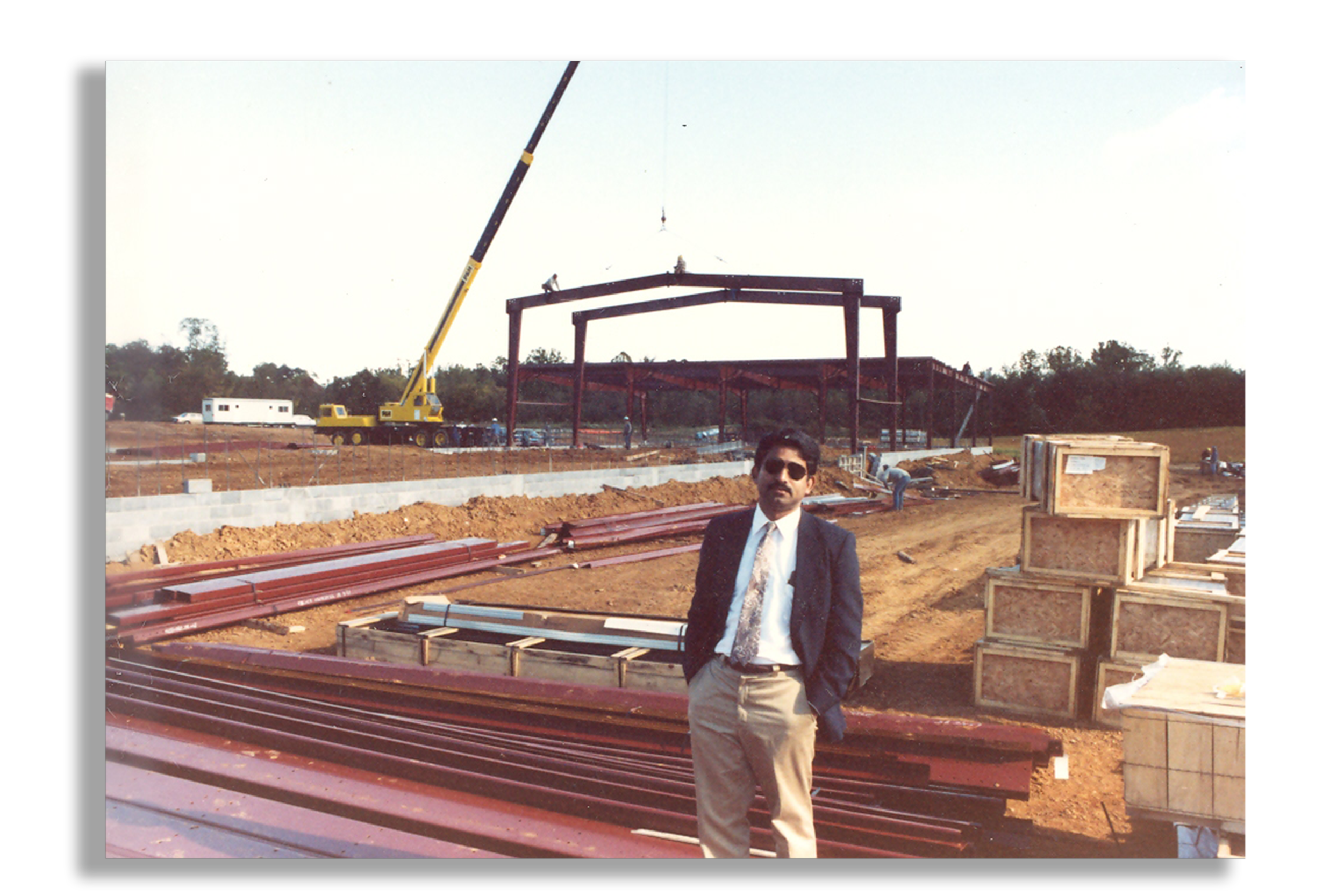 Man in a suit and sunglasses standing on a construction site with steel beams and building materials, with a large crane in the background.