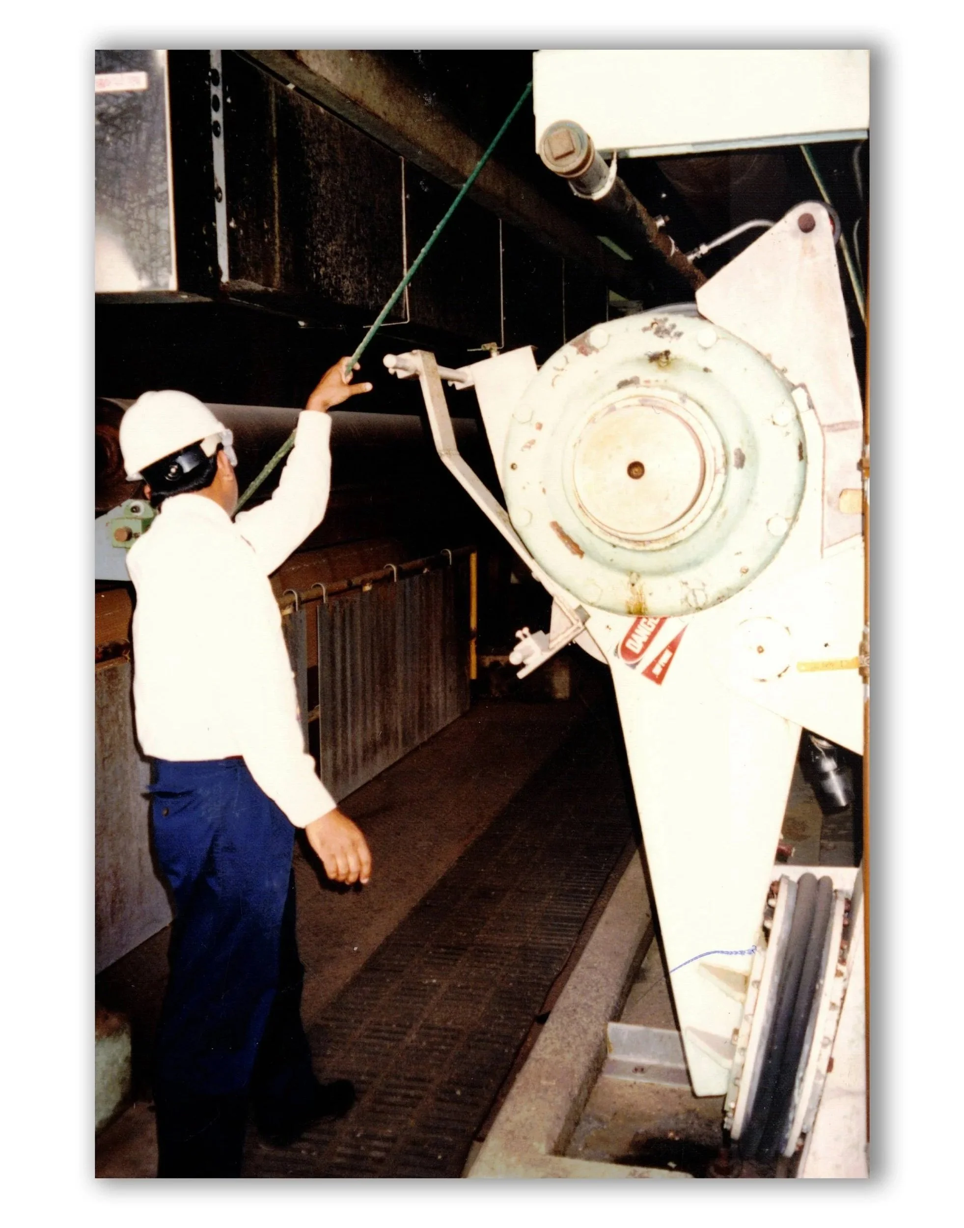 A worker in a hard hat and safety glasses inspects or maintains a large industrial machine using a long tool in a facility with metal floors and equipment.