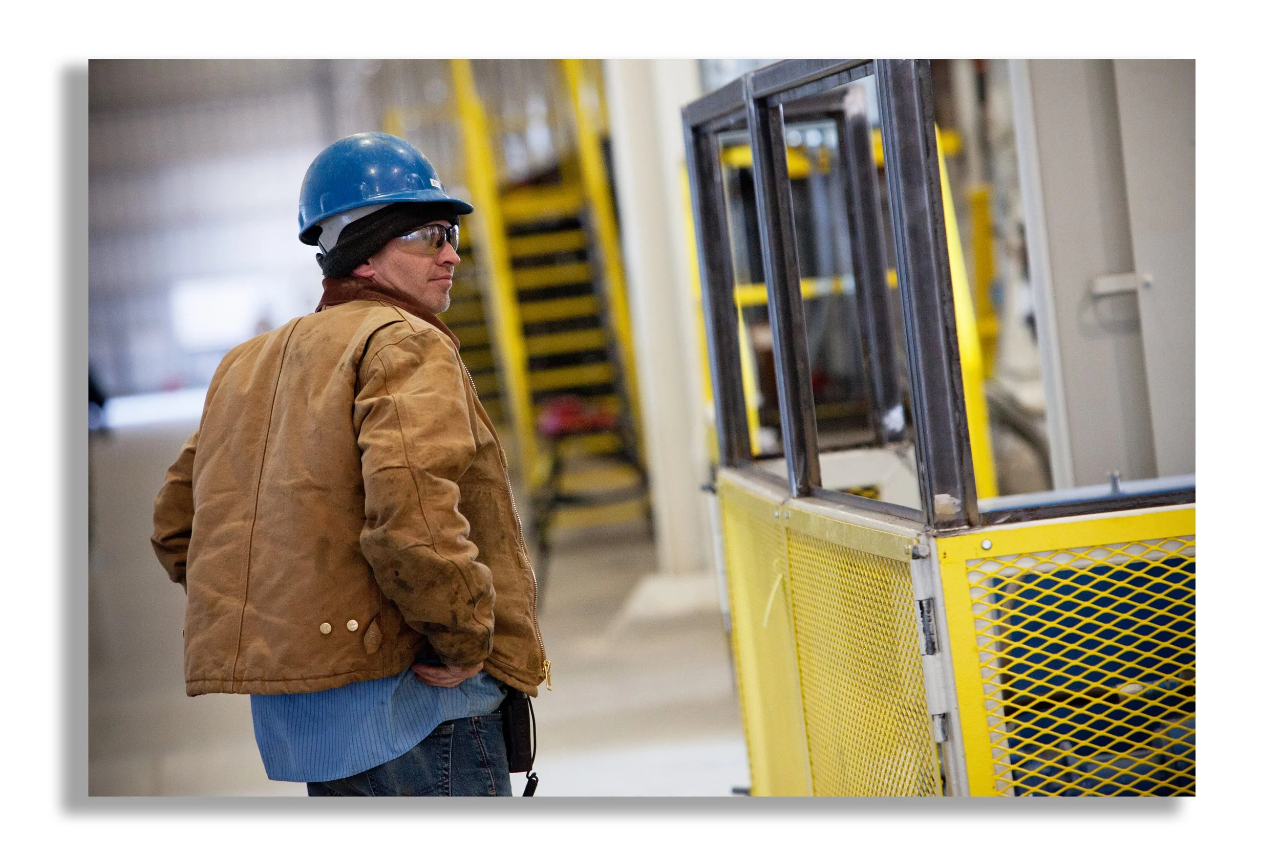 A man wearing a blue hard hat, safety glasses, a brown jacket, and jeans stands with his hands in his pockets inside an industrial setting, looking at yellow safety barriers.