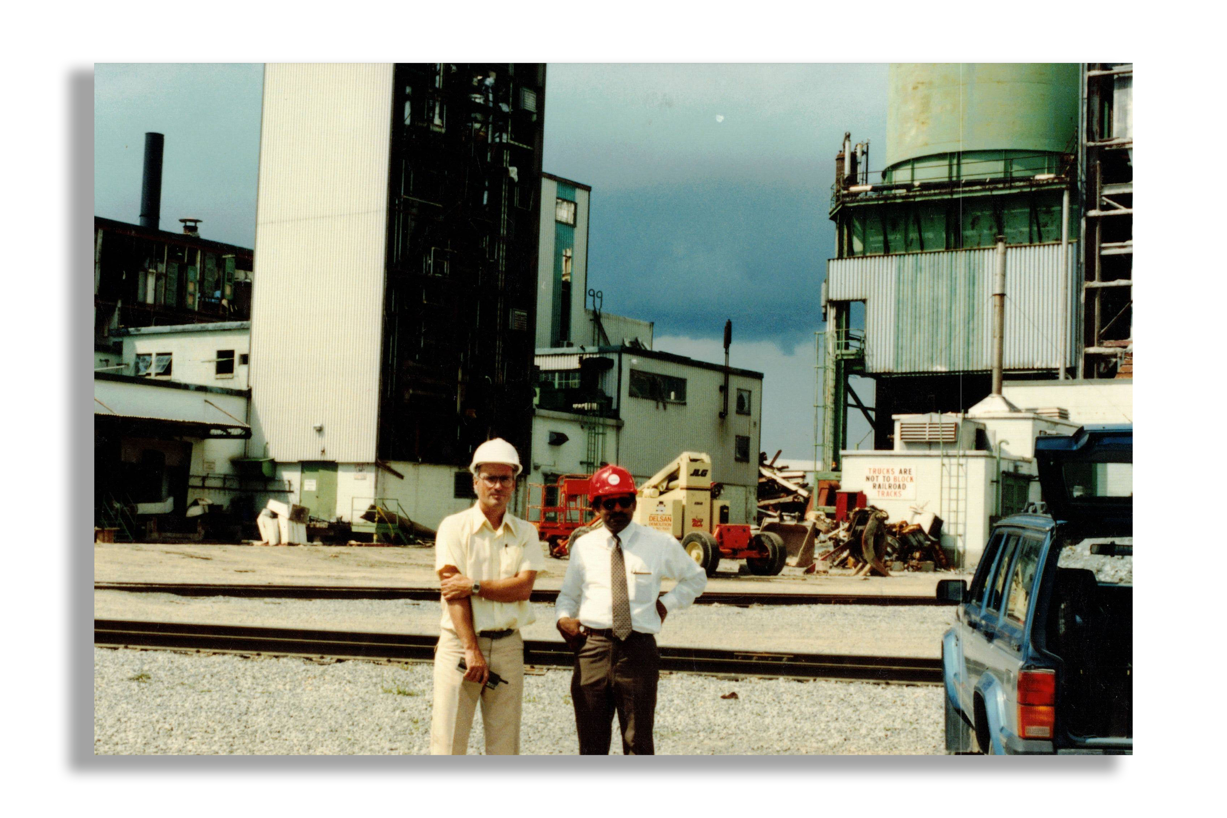 Two men wearing safety helmets standing in front of an industrial facility with machinery, structures, and railroad tracks, under a cloudy sky.