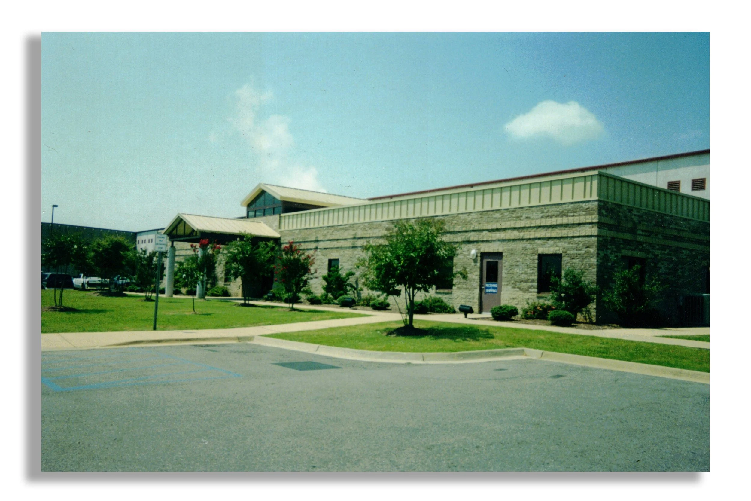A single-story brick building with a green metal roof, surrounded by small trees and shrubs, in a parking lot with marked spaces. The sky is blue with a few clouds.