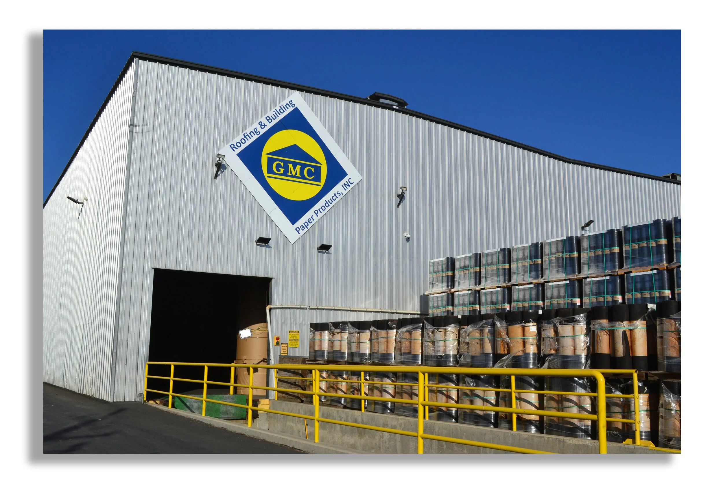 Exterior view of a warehouse with a GMC sign indicating roofing and building paper products, with stacks of rolled roofing materials outside and a yellow safety railing in front.
