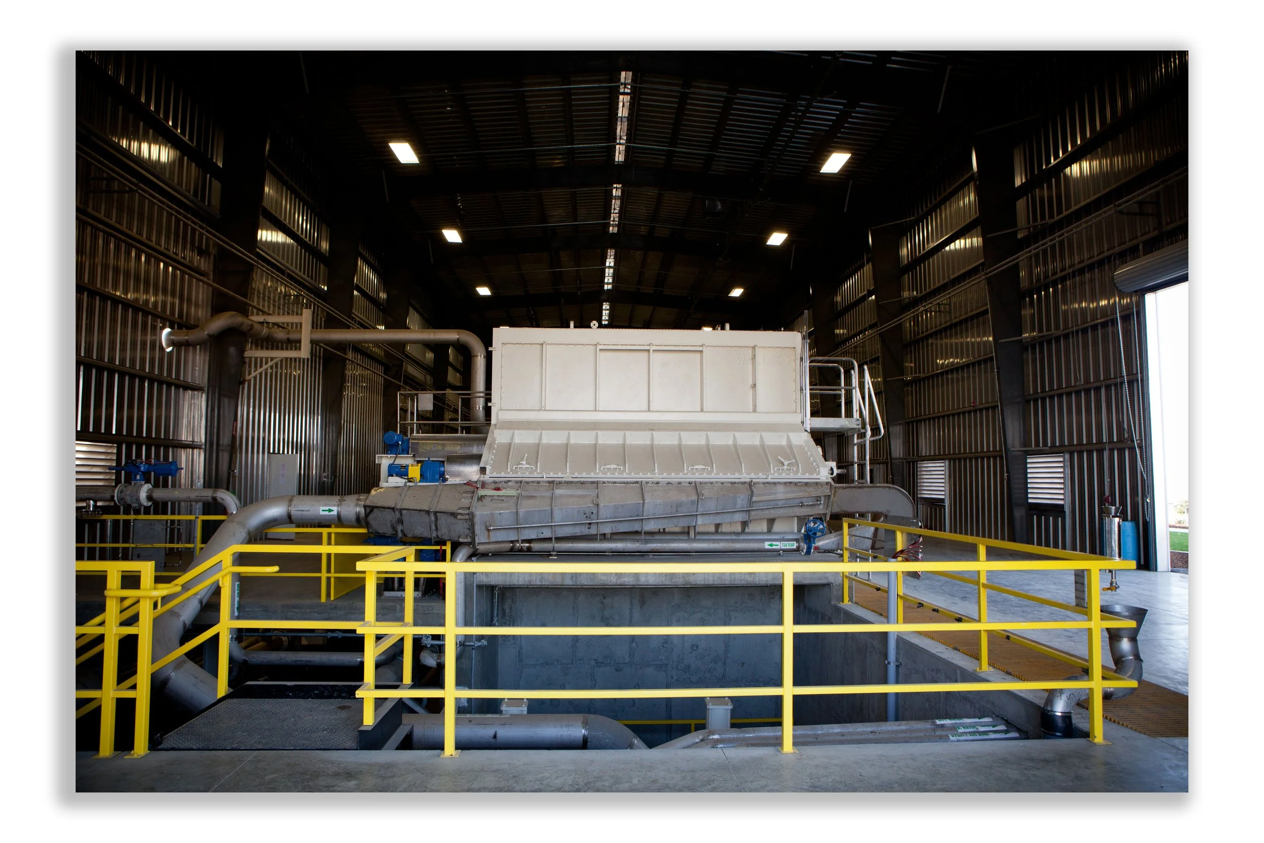 Industrial machinery inside a large warehouse or factory, with pipes, metal structure, and yellow safety railing.