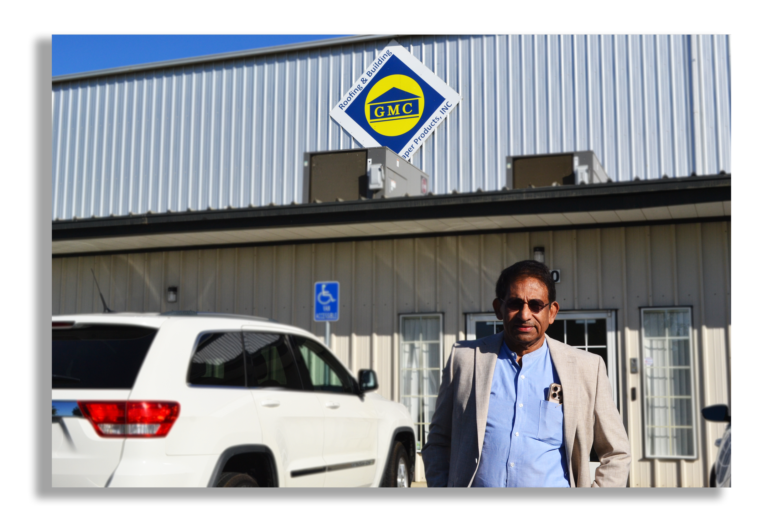 A man in a blazer and sunglasses standing outside a building with a GMC Roofing & Building sign. A white SUV is parked nearby, and a handicap parking sign is visible.