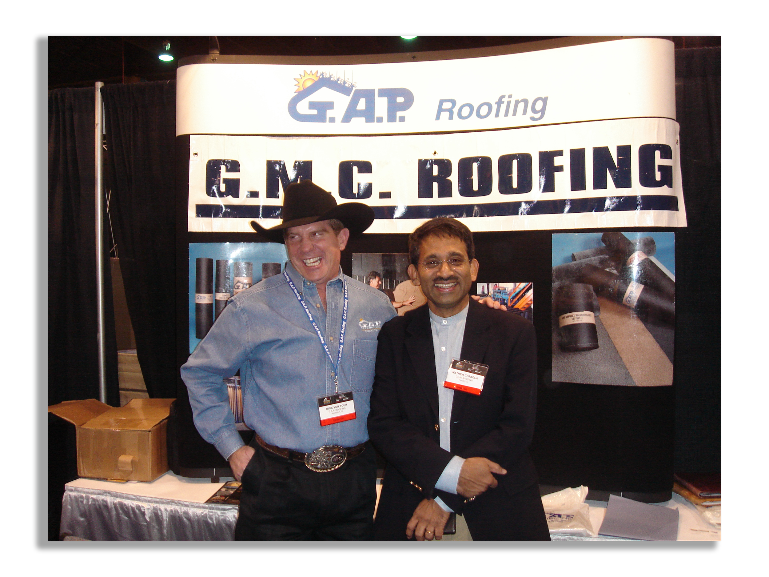 Two men standing in front of a booth with G.A.P Roofing signs. One man is wearing a cowboy hat and blue shirt, and the other is wearing a black blazer with a name tag. They are smiling.