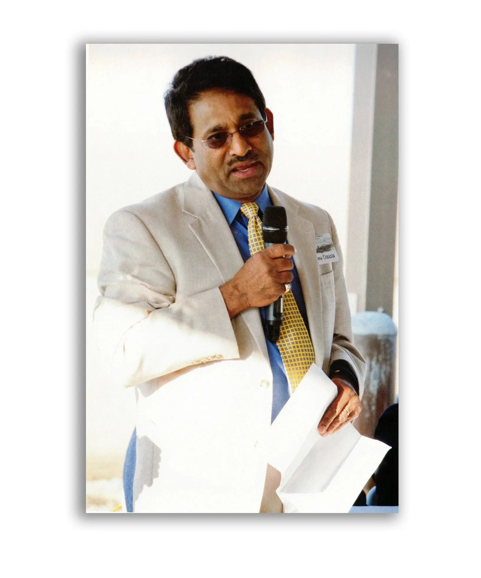 A man in a beige suit with glasses, holding a microphone, speaking at an event.