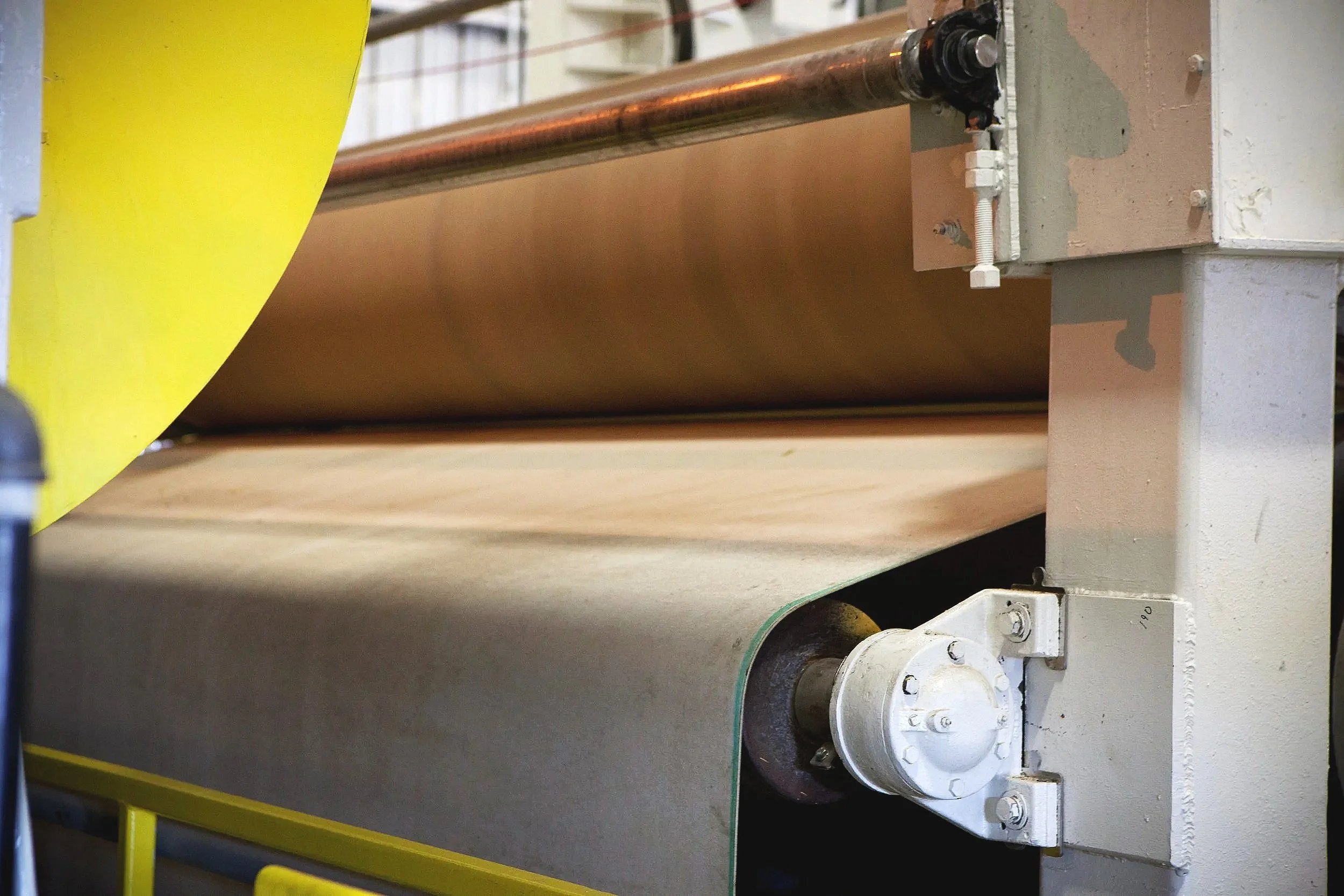 Close-up of industrial machinery with a large metal roll and mechanical parts, including a white gear mechanism and a horizontal copper pipe, in a manufacturing or processing plant.