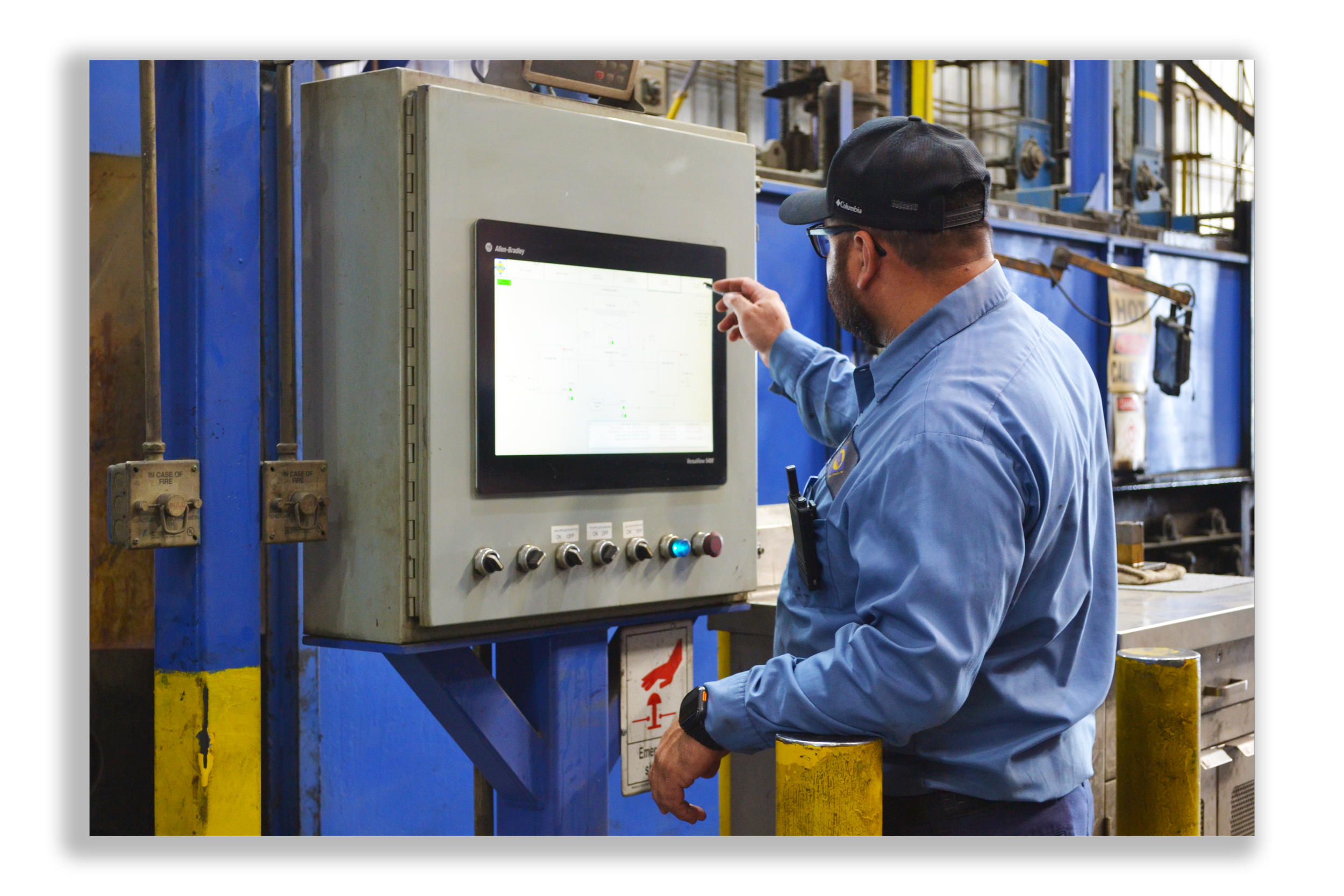 A man wearing a blue shirt, black cap, and safety glasses is operating a control panel in an industrial setting. He is pointing at the touchscreen display on the control panel.