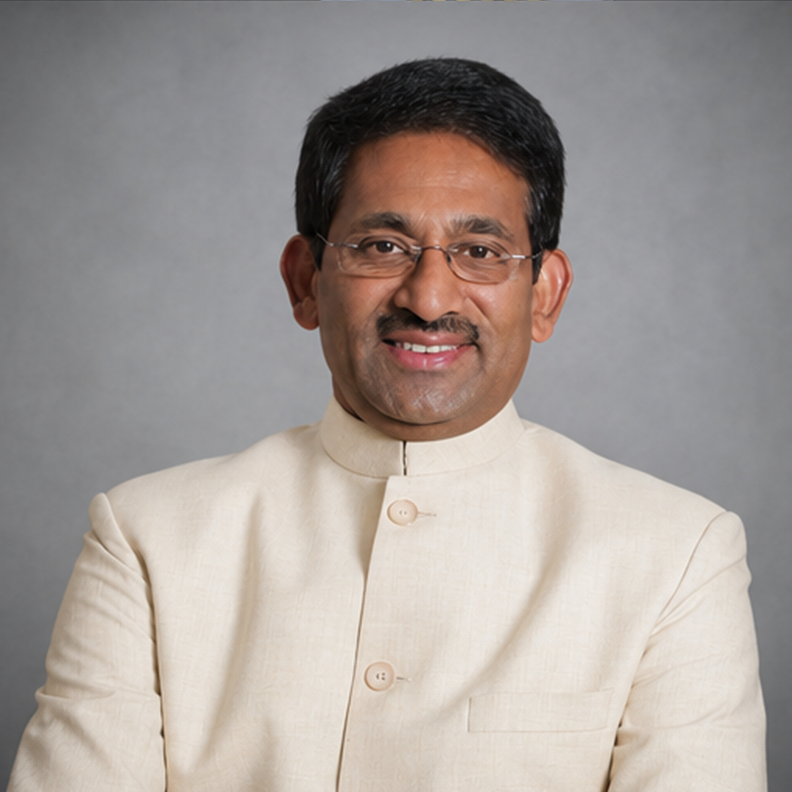 A man wearing traditional Indian attire standing with crossed arms in front of a bookshelf and wooden cabinet in a study or office.