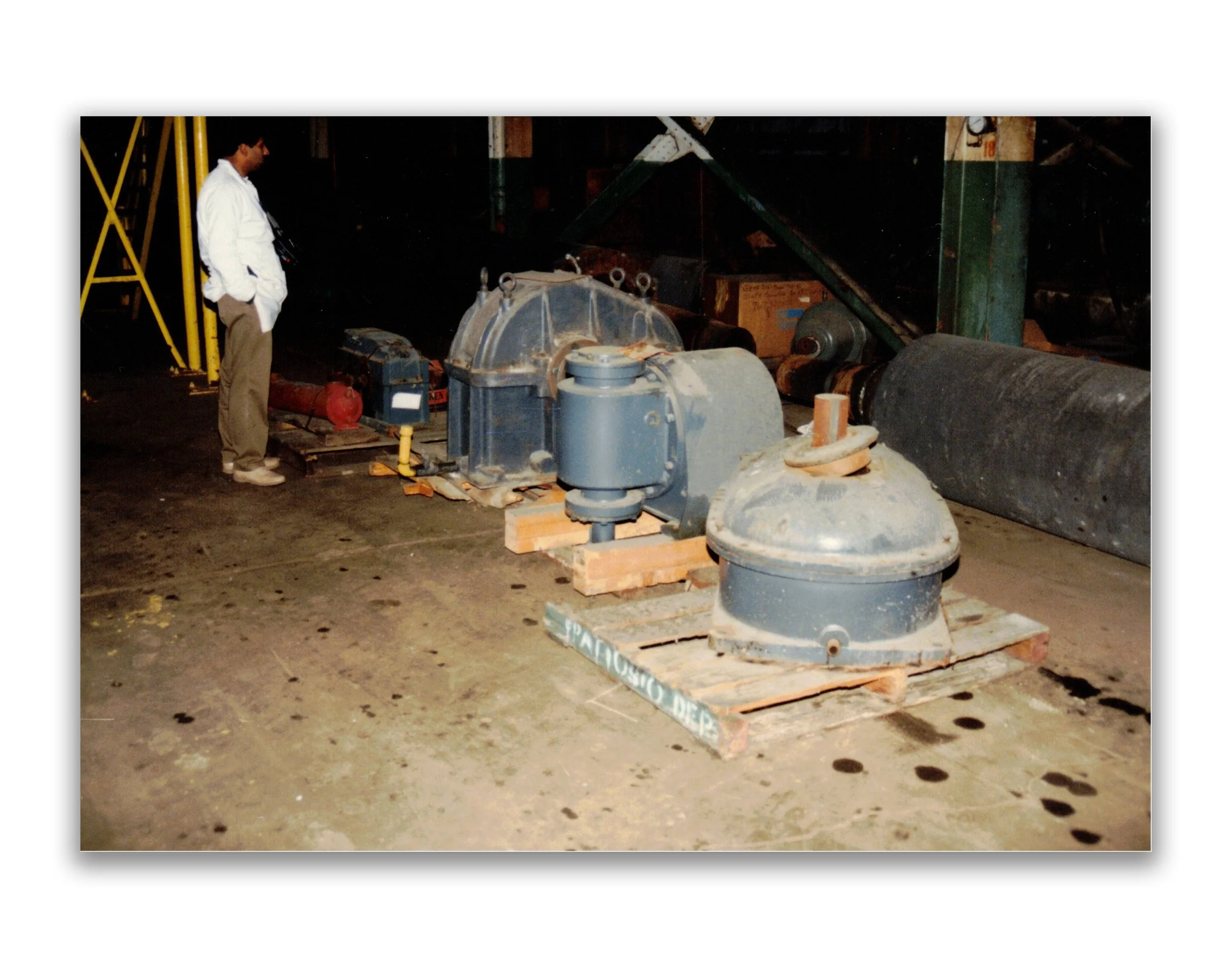 A man wearing a white shirt and beige pants is standing and observing large industrial equipment in a warehouse. The equipment is placed on wooden pallets on a concrete floor.