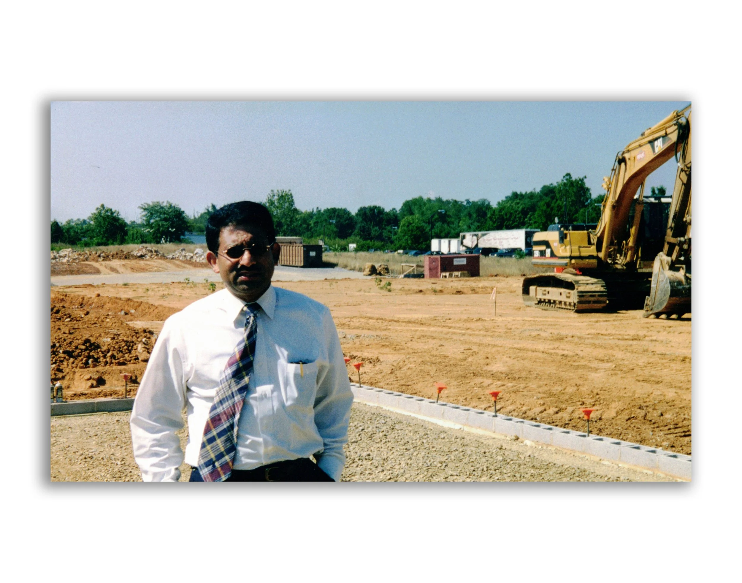Man wearing sunglasses, white dress shirt, and plaid tie standing at a construction site with heavy machinery and excavated dirt in the background.