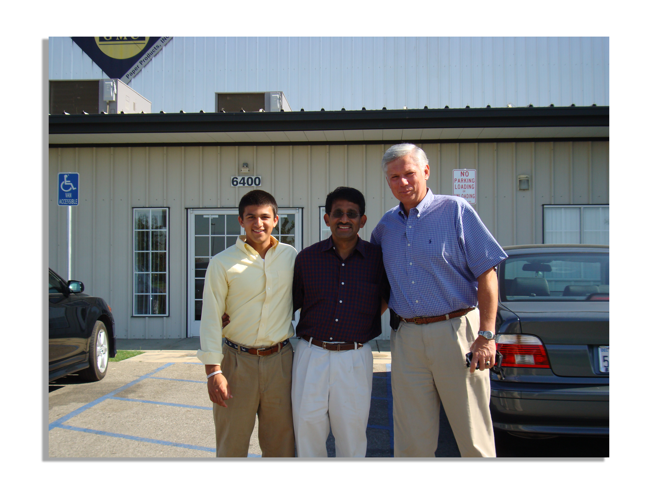 Three men standing outside in front of a building with a parking lot, smiling at the camera.