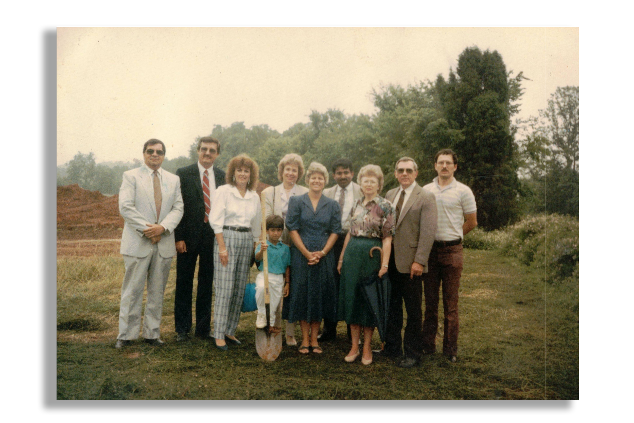Group of ten people, including a woman holding a cane and a child, standing outdoors in front of trees and bushes, with a dirt mound in the background, possibly on a farm or rural area, during daytime.