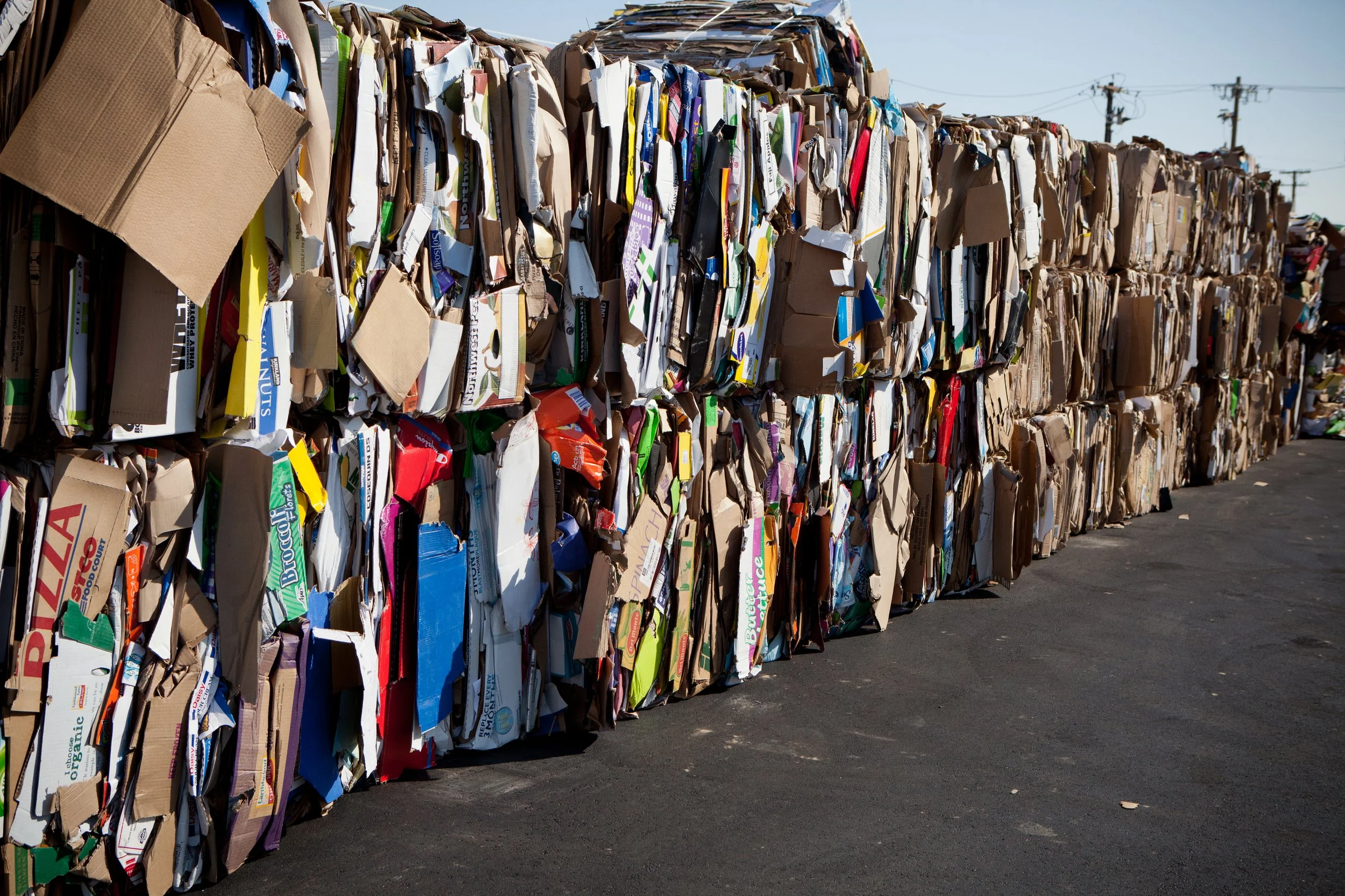 Large stacks of flattened cardboard boxes placed outdoors on an asphalt surface, with power lines in the background.