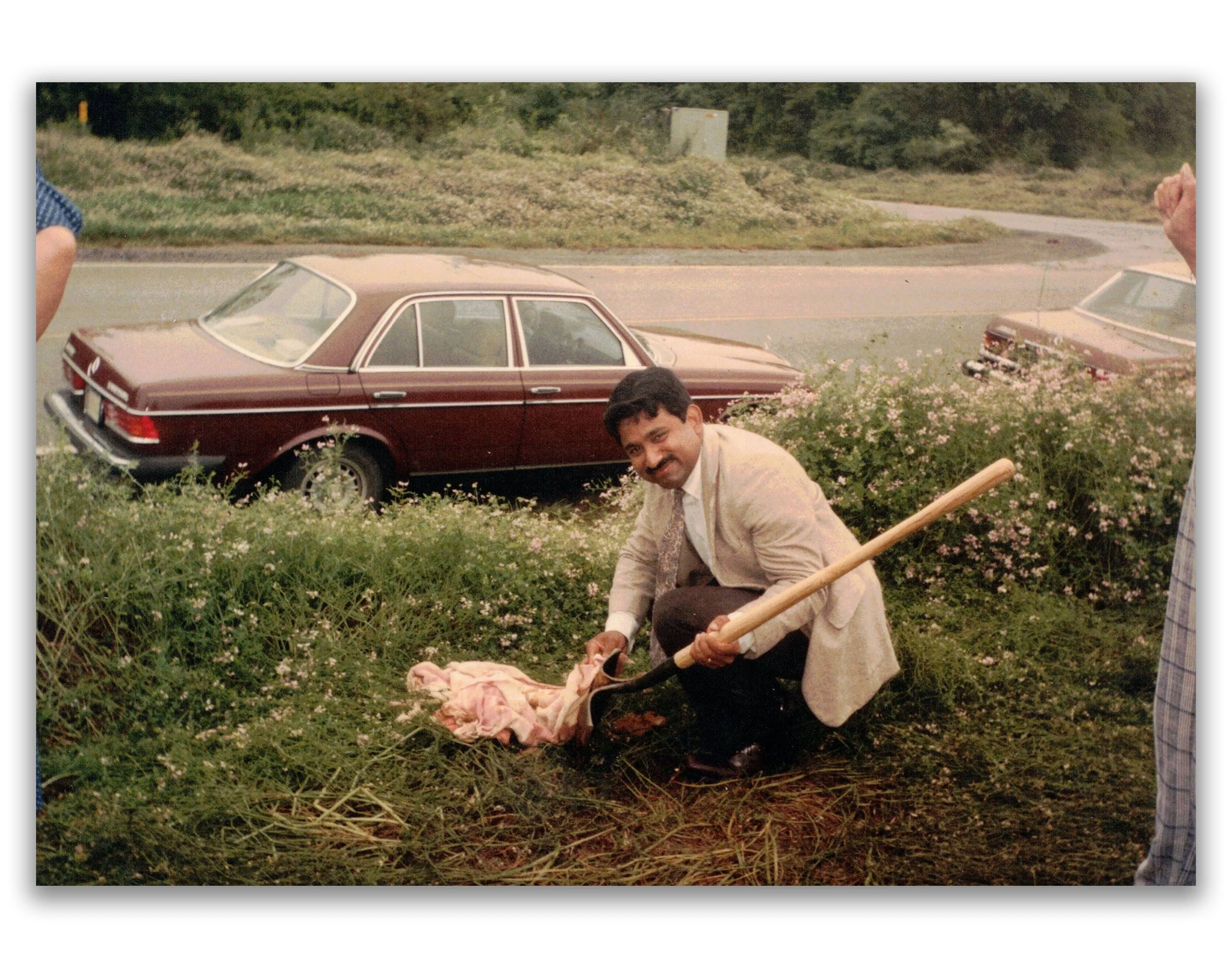 A man with a mustache, wearing a beige blazer and dark pants, is kneeling outdoors and digging with a shovel. There are cars parked on the side of a road behind him, and flowering bushes surround him.