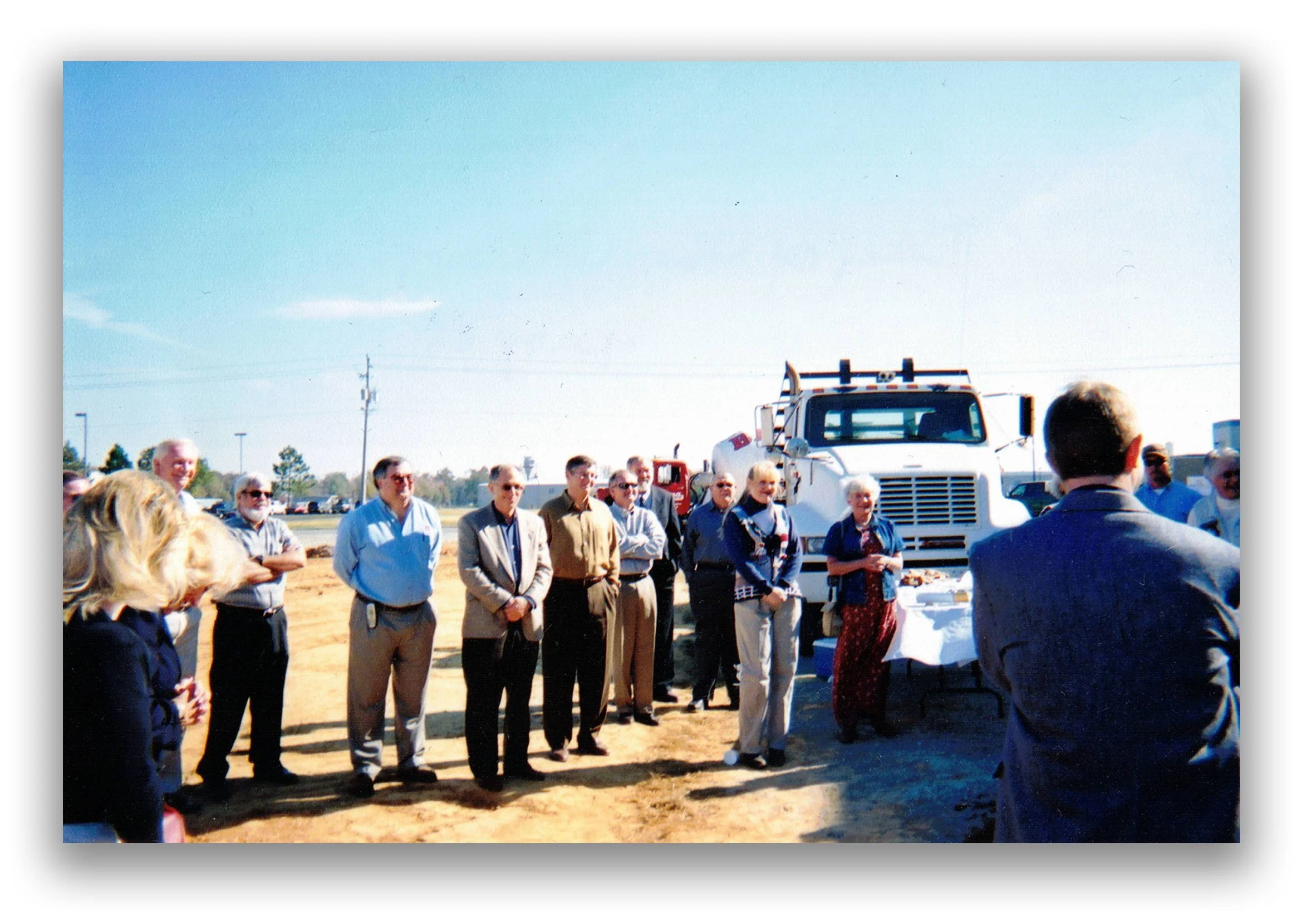 Group of people attending an outdoor event near a white truck, with a table of food and drinks, on sunny day, in a mostly clear sky.