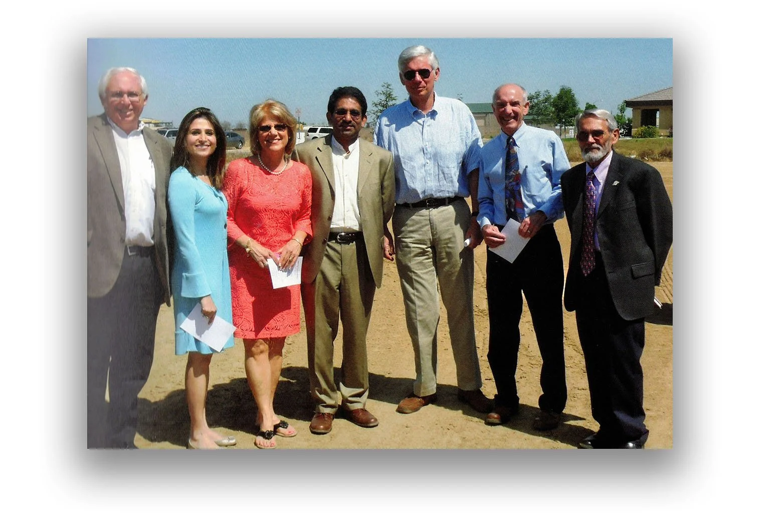 Group of eight people standing outdoors on a dirt area with buildings and trees in the background, dressed in business casual attire, some holding papers.