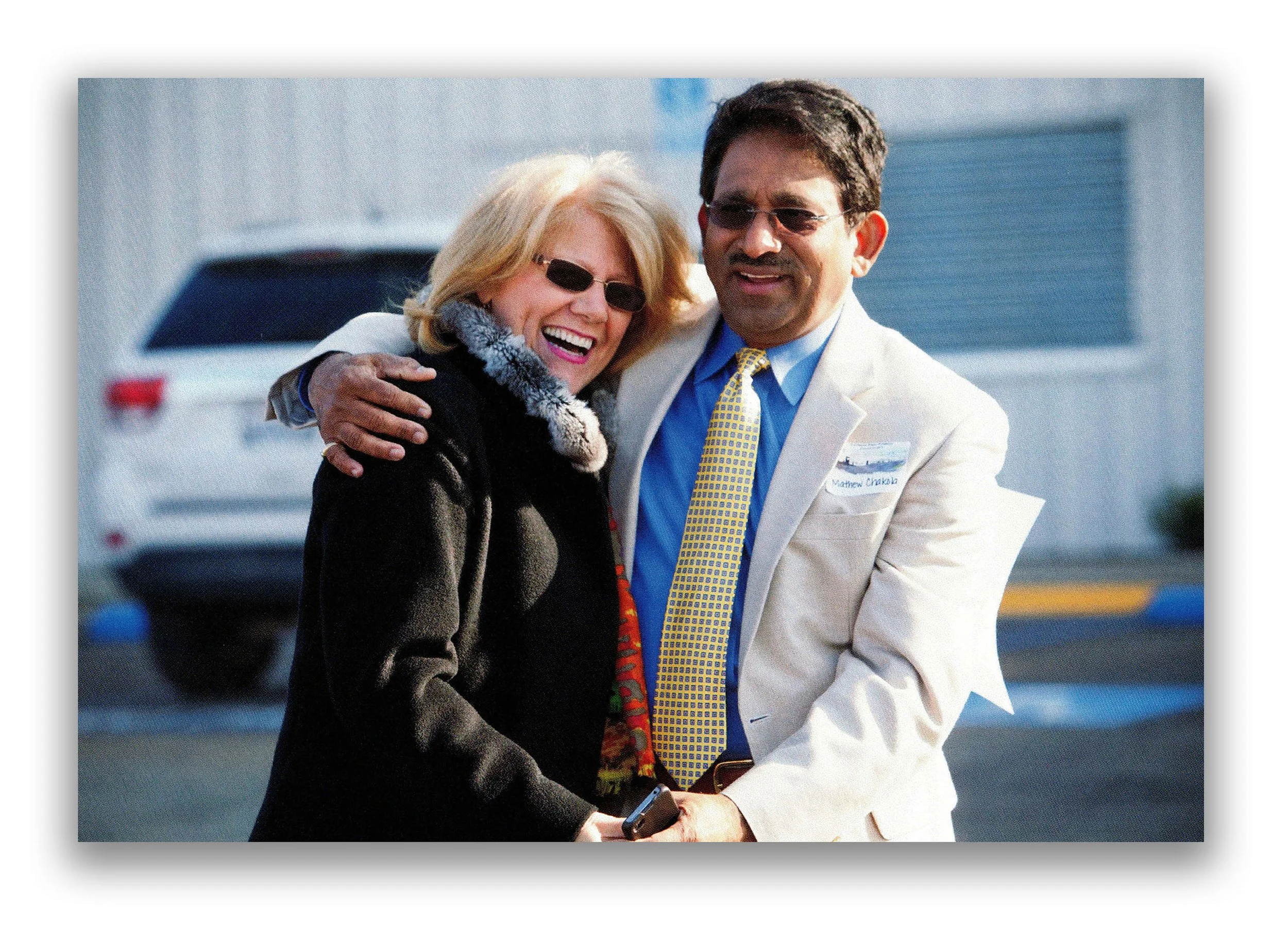A man and a woman smiling and hugging outdoors, with a parking lot and building in the background.