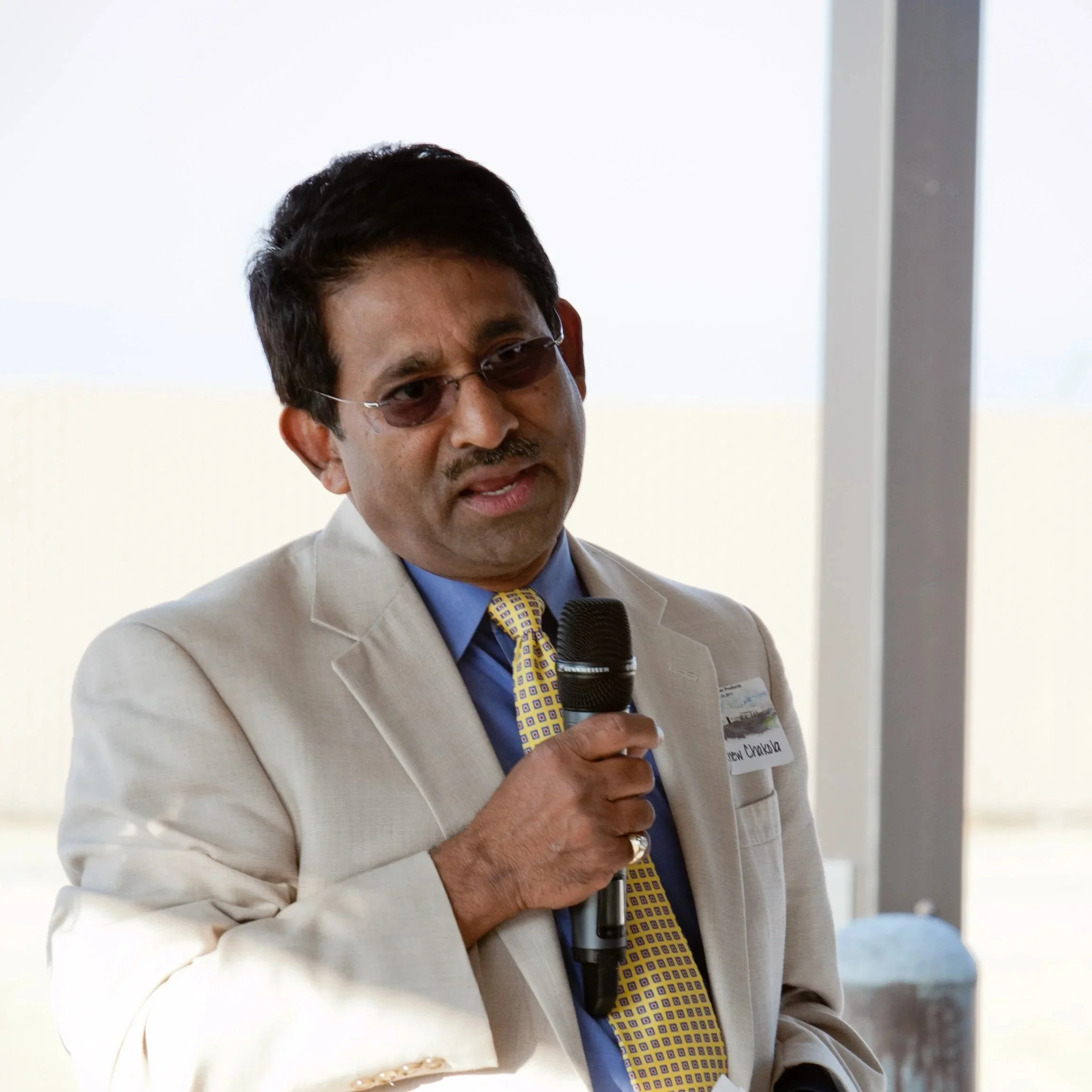 Man in beige suit with yellow patterned tie and tinted glasses speaking into a microphone.