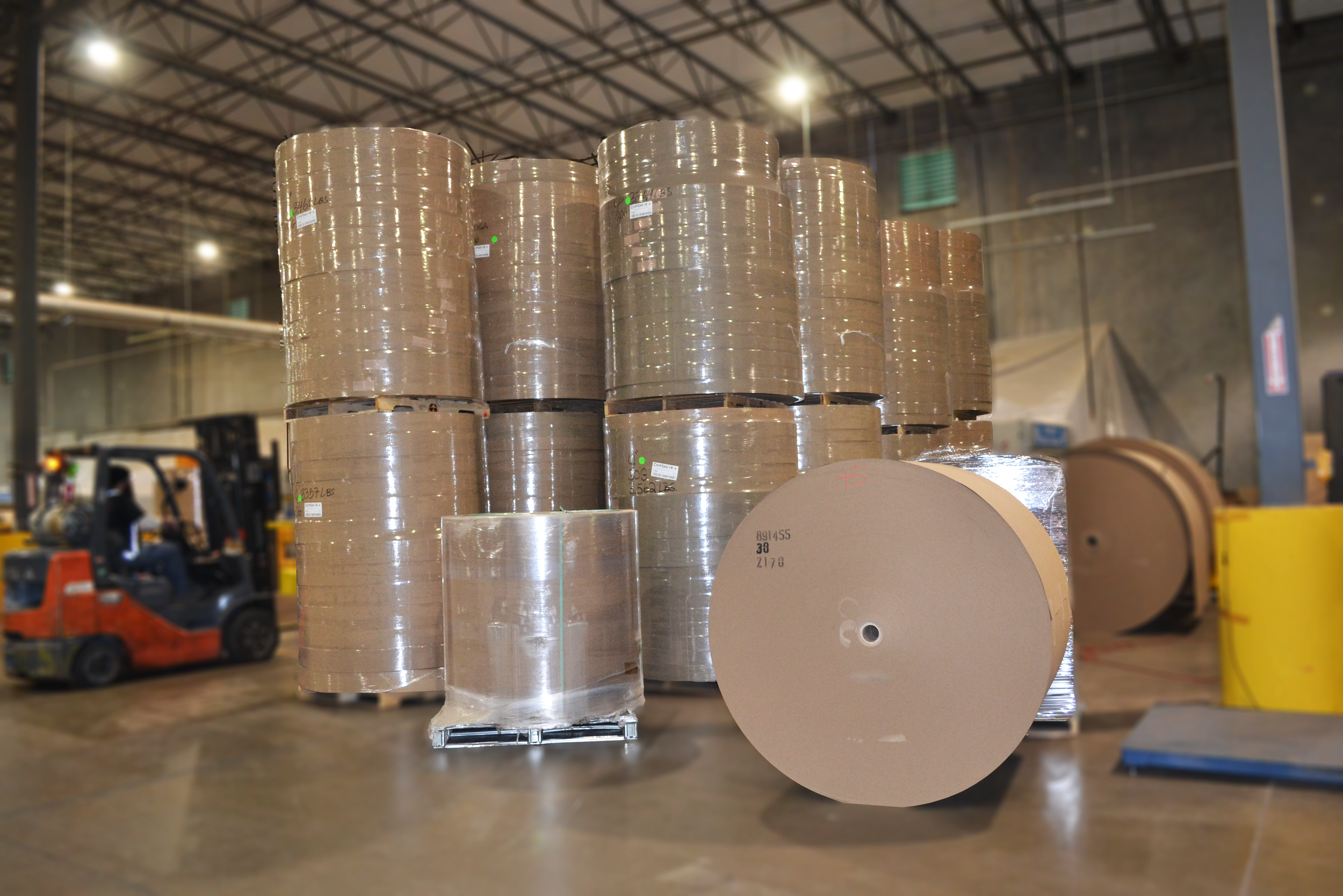 Stacks of large packing tape rolls in a warehouse, with a forklift and industrial setting in the background.