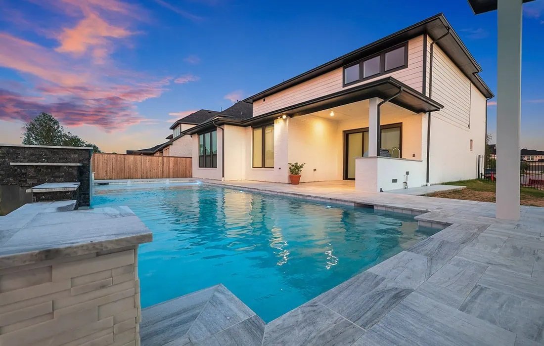 Modern house with a backyard swimming pool during sunset, with a patio, patio furniture, and a wooden fence.