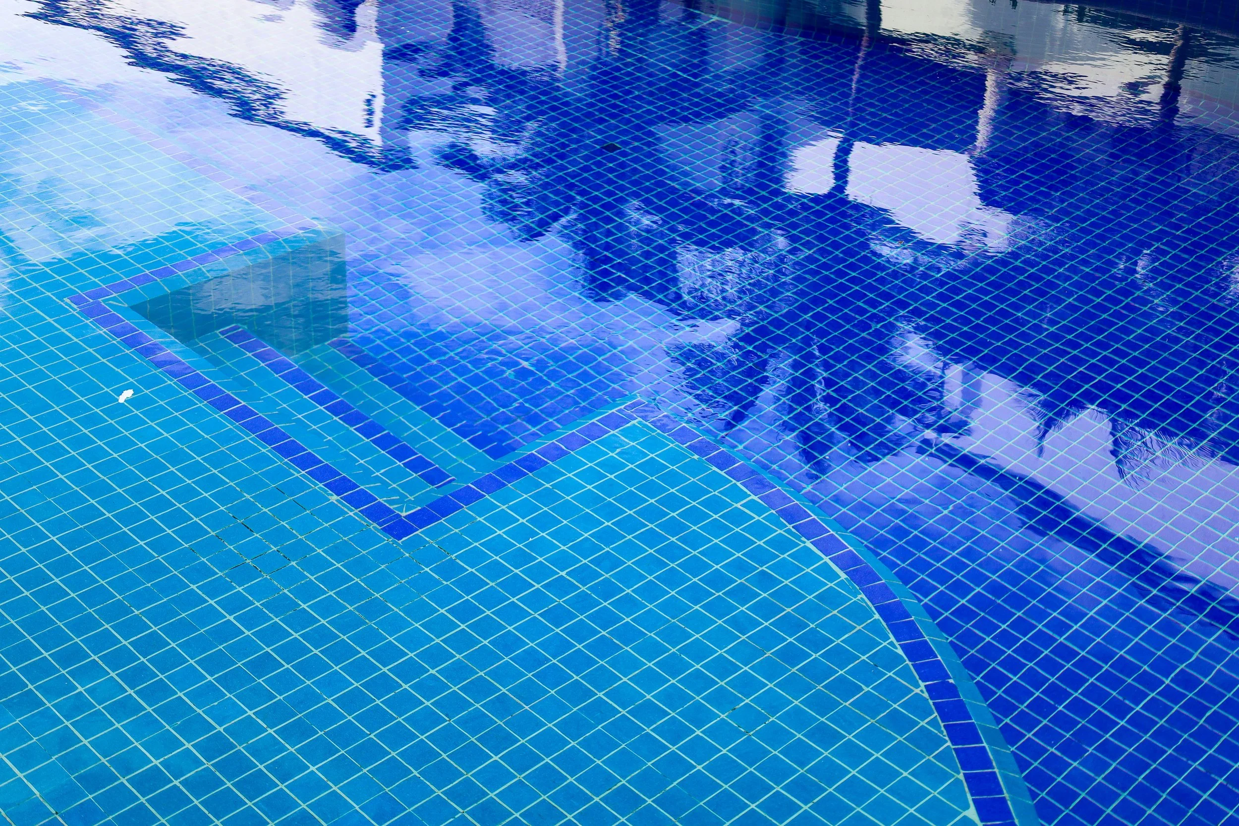 Close-up view of a swimming pool with blue tiles, showing the reflection of the sky and nearby trees on the water surface.