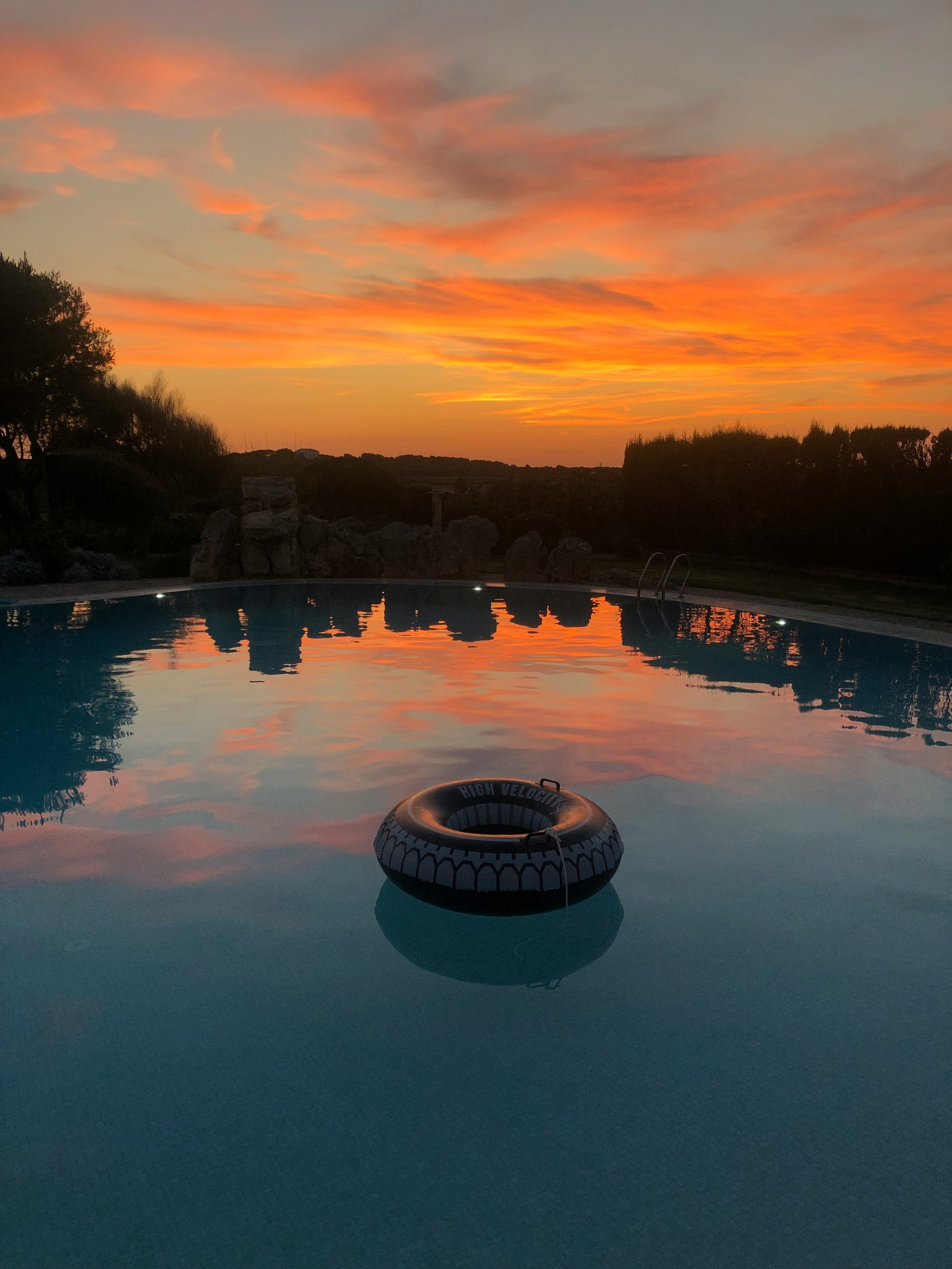 A swimming pool reflecting a vibrant sunset sky with orange and pink clouds, surrounded by rocks and trees in the distance.