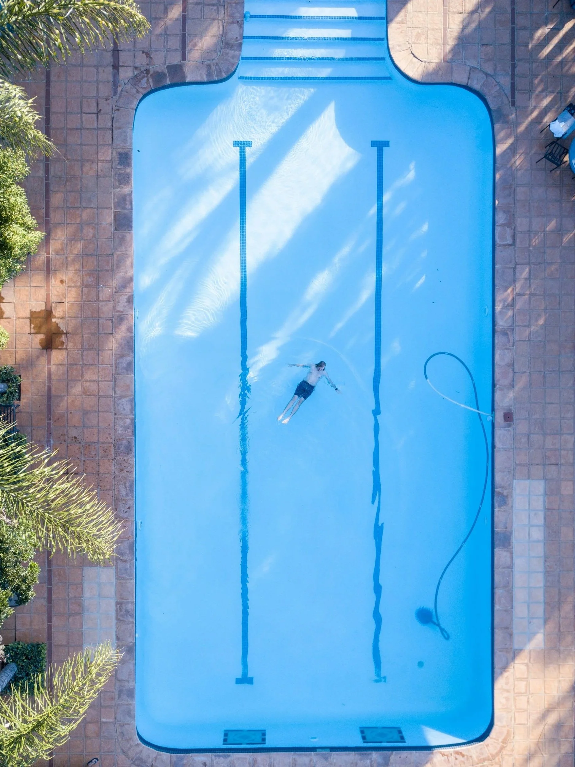 An aerial view of a swimming pool with a person swimming in the middle. The pool has two lanes marked with blue lines. The area around the pool is tiled, and there are trees on the left side of the image.