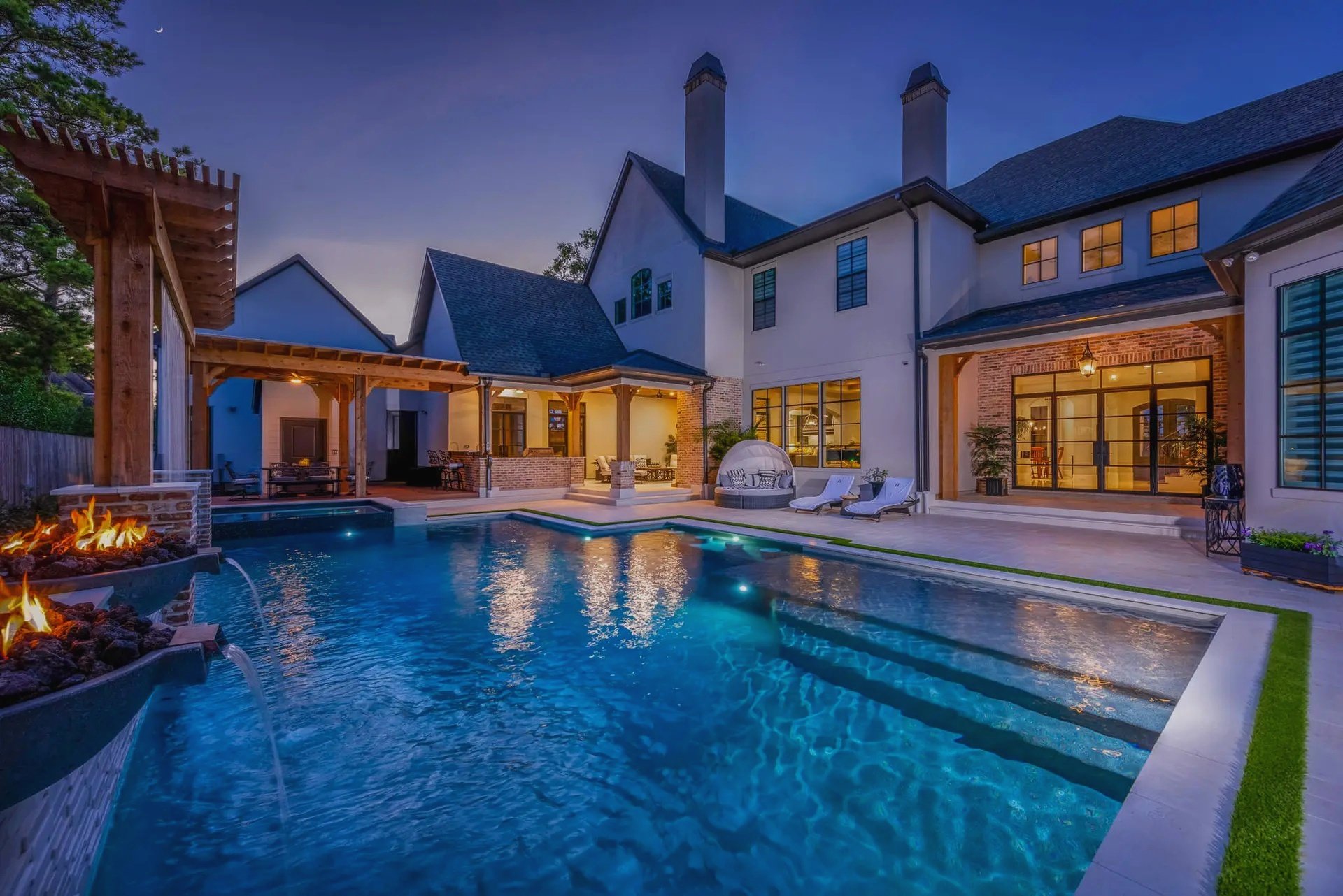 Large modern house with a backyard pool at dusk, illuminated with lighting, featuring outdoor lounge chairs, a fire feature, and multiple windows.