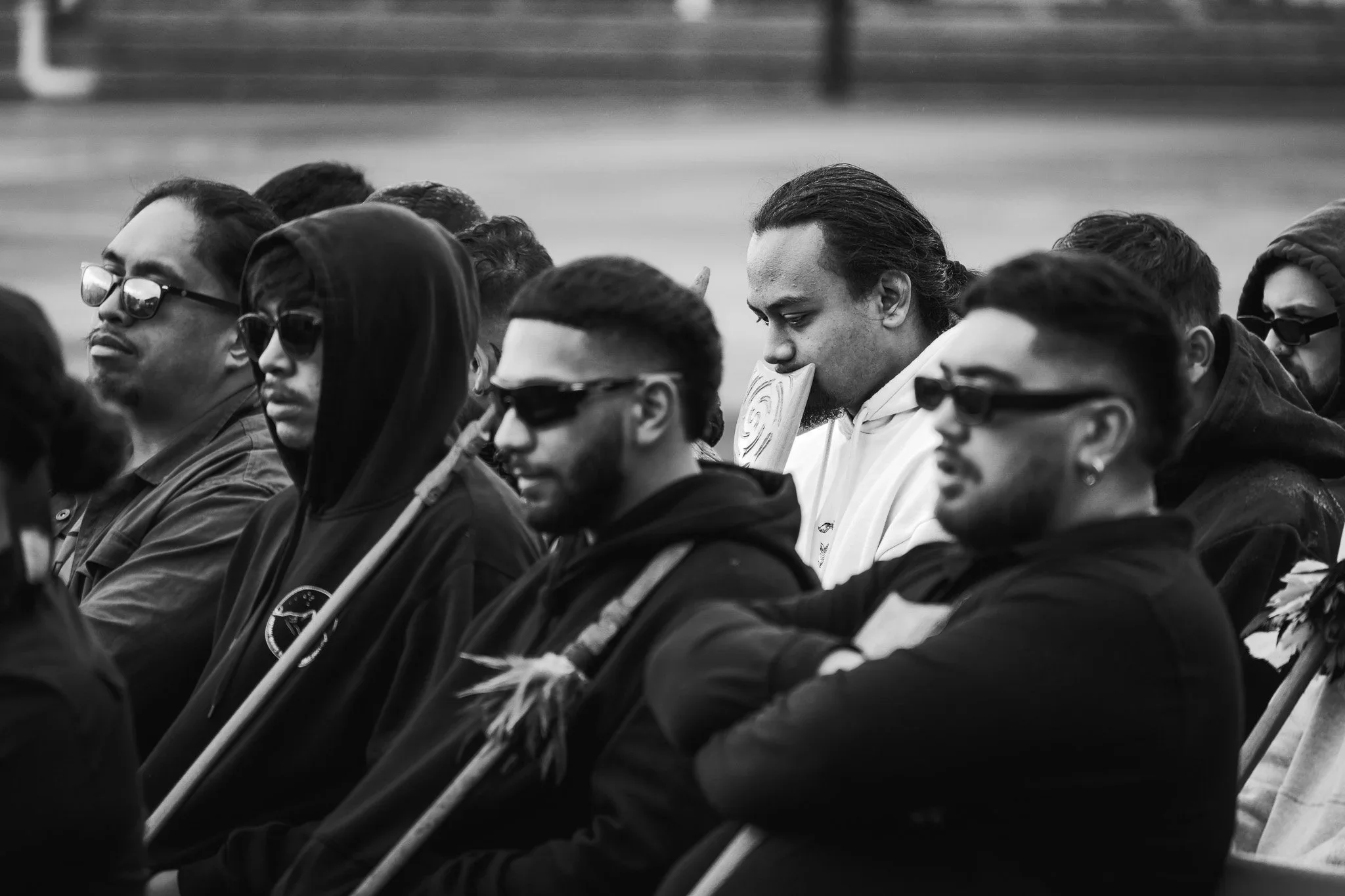 Group of people sitting outdoors, some wearing sunglasses, with one person holding a sign with a spiral design and another holding a pole with feathers.