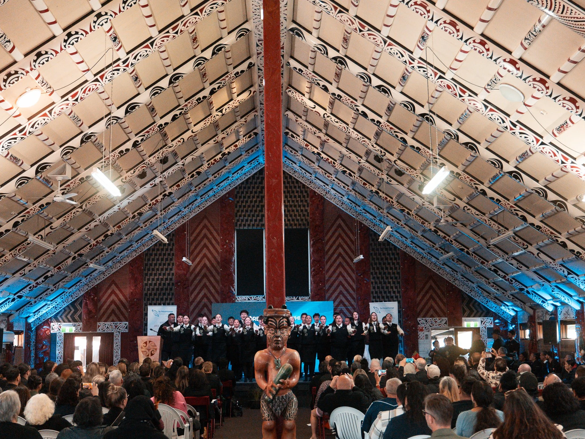 Indigenous choir performing on stage inside a large hall with intricate roof design, an audience seated in front, and a person in traditional attire standing in the foreground.
