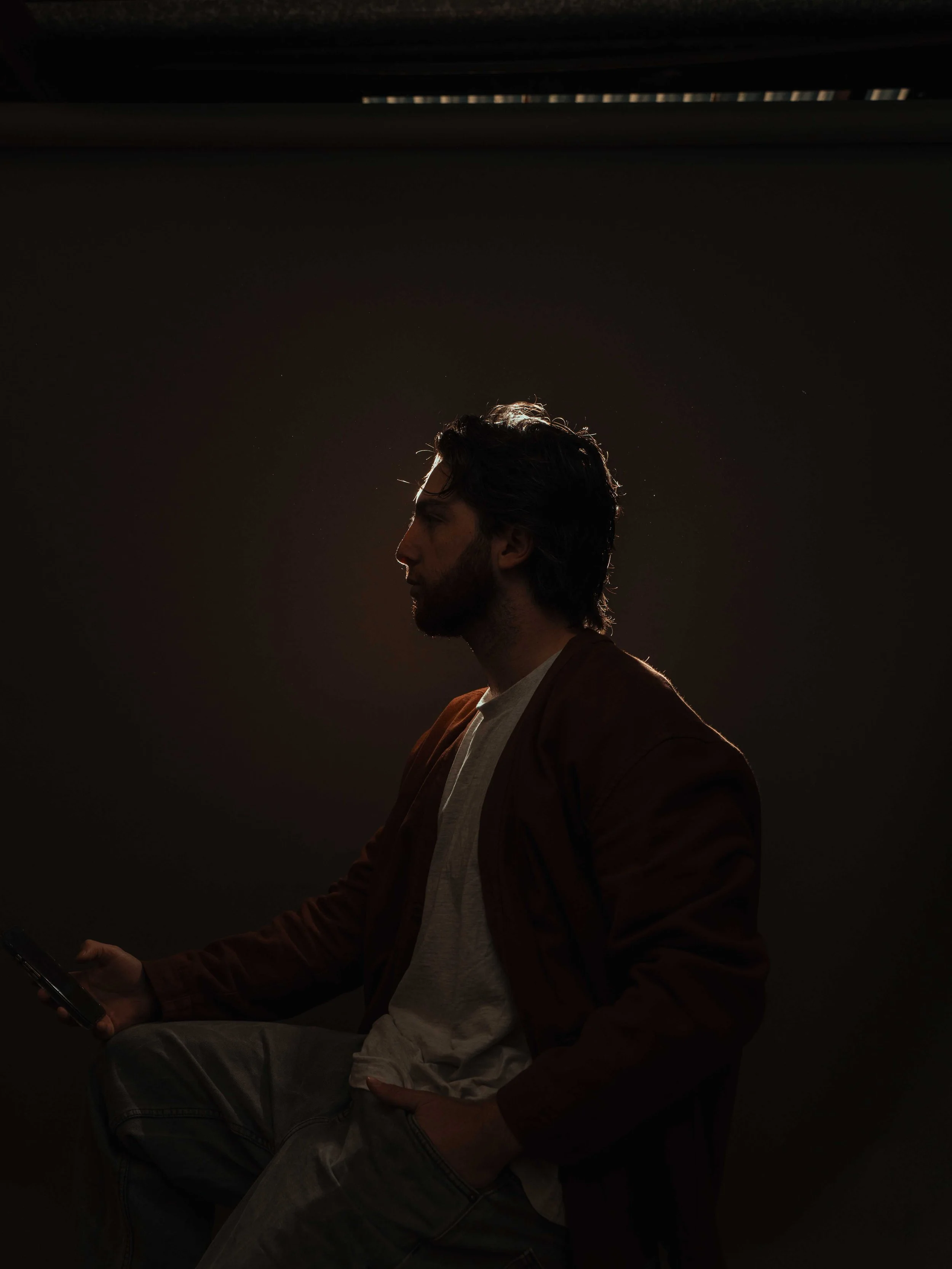 A silhouette of a man with dark, wavy hair, sitting against a dark background, holding a smartphone in his right hand, with light outlining his profile.