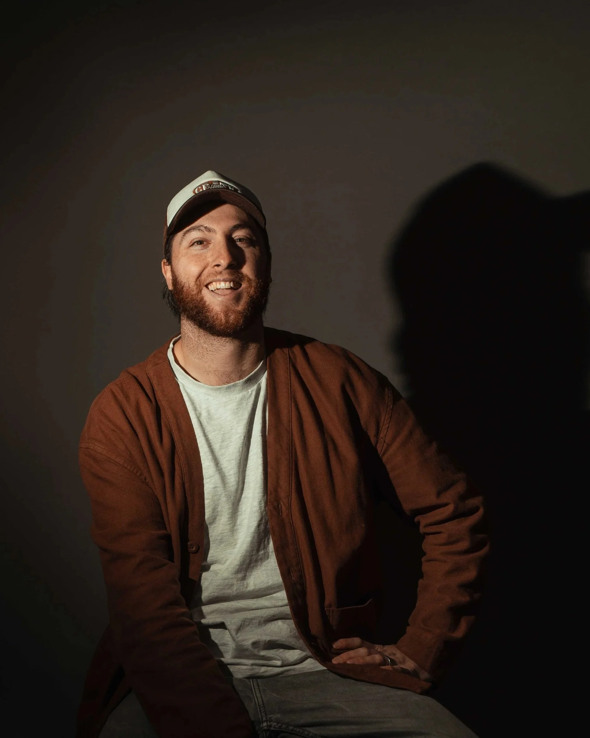 A smiling man with a beard wearing a white cap, white shirt, and brown jacket, sitting against a plain dark background.
