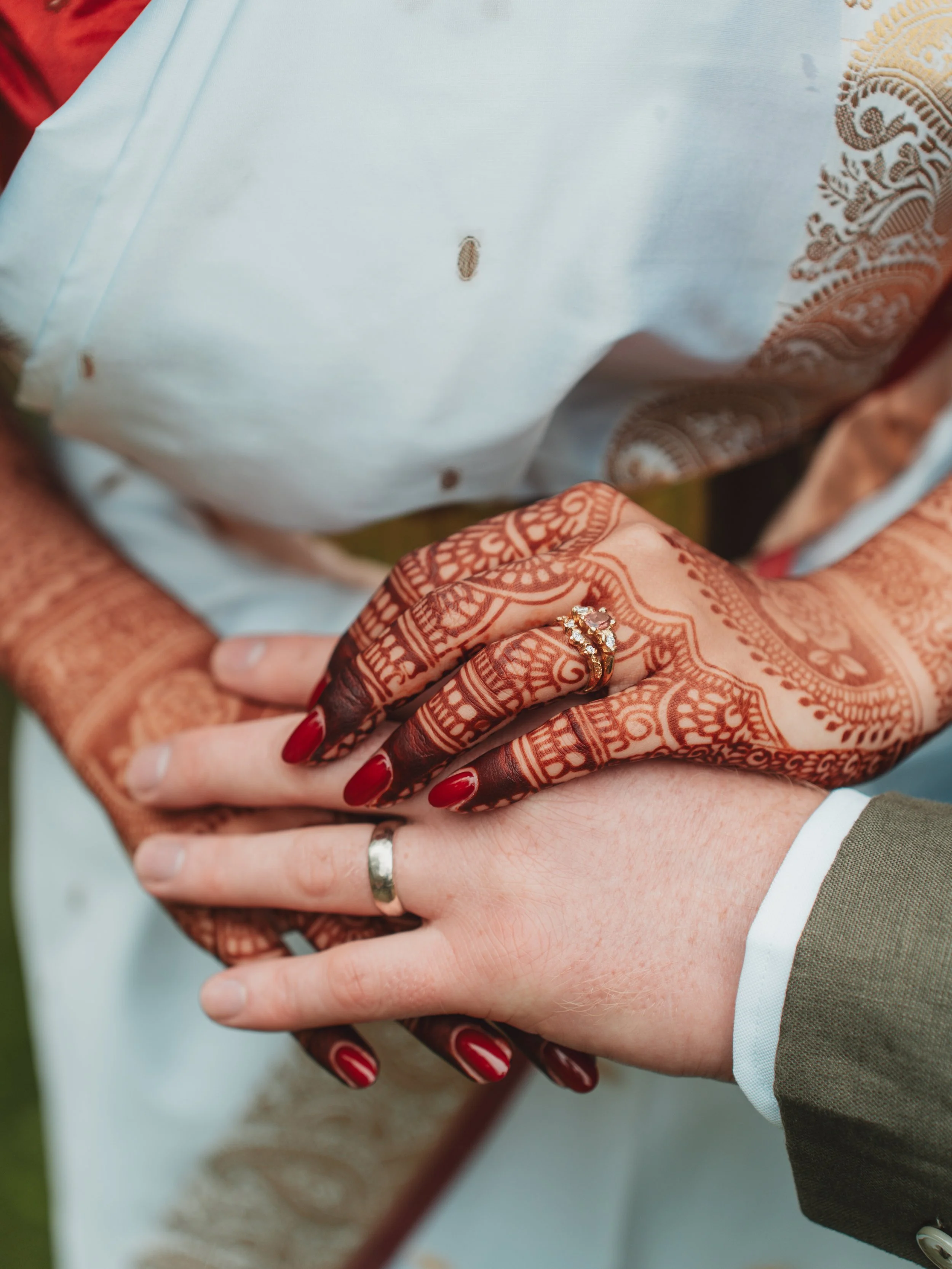 Close-up of a bride and groom holding hands, showing wedding rings and intricate henna designs on the bride's hand.