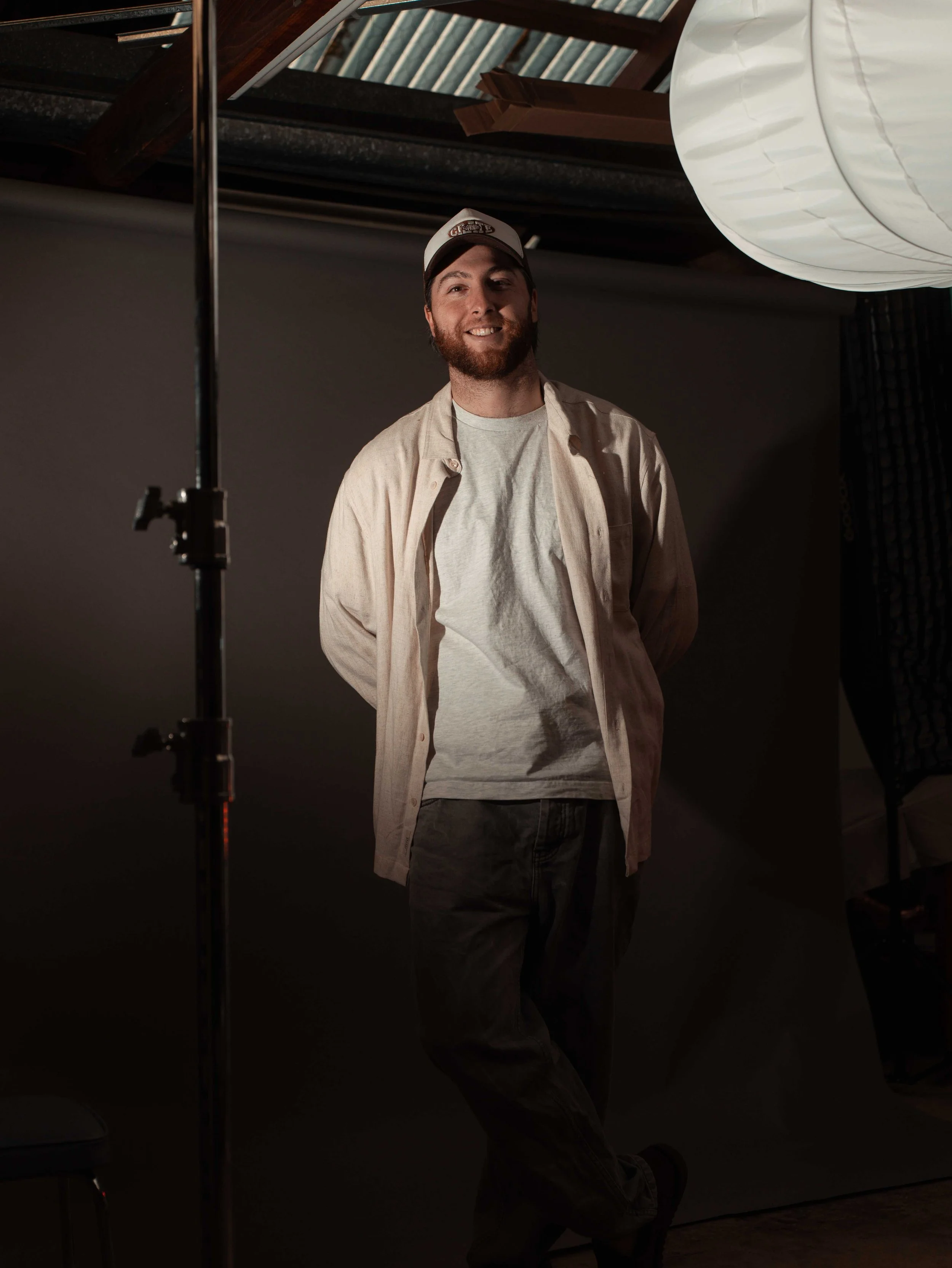 A smiling man with a beard wearing a beige shirt, gray t-shirt, and a baseball cap standing in a photography studio with studio lights and equipment.